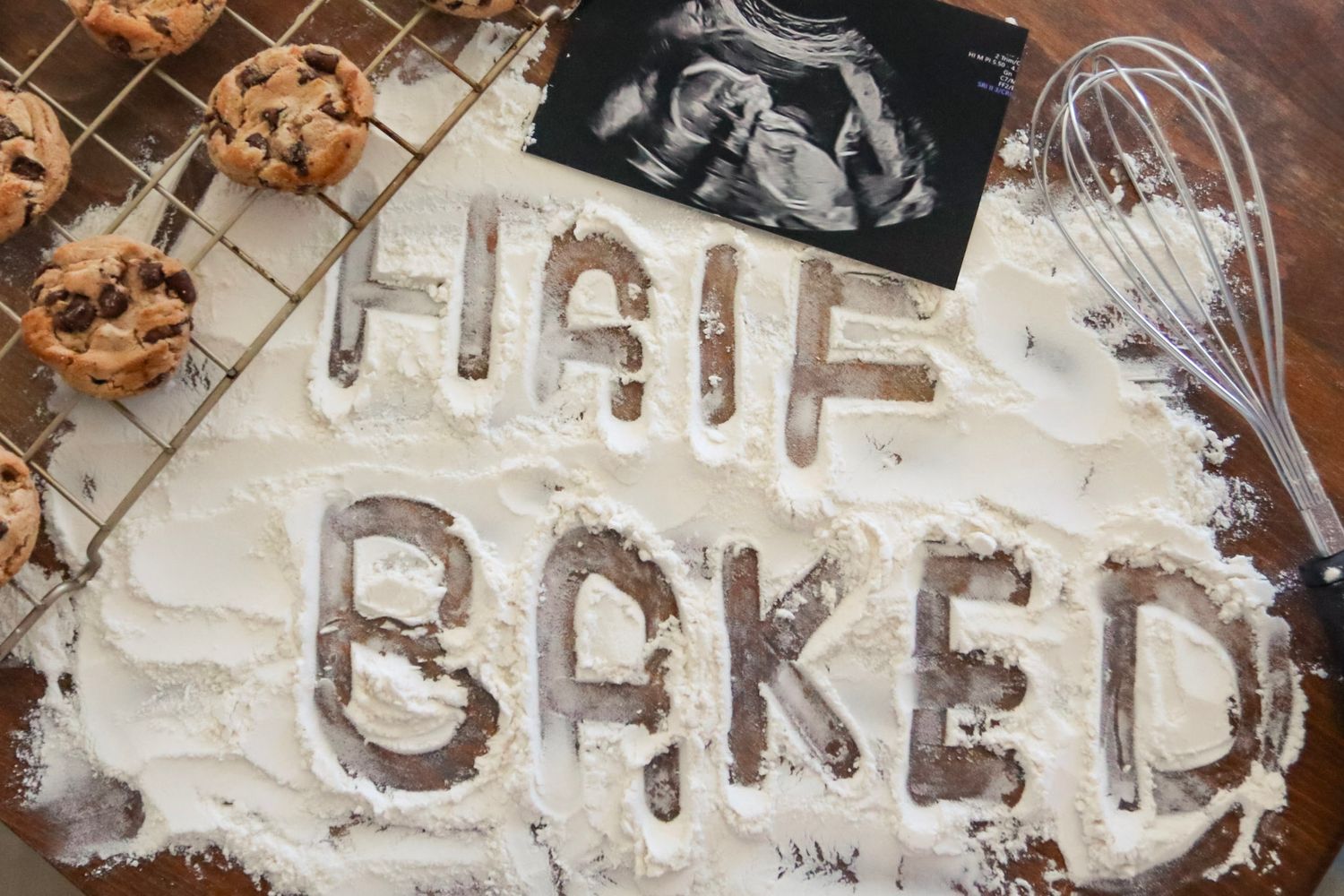 Flour spelling 'Half Baked' with ultrasound photo, cookies, and whisk for creative pregnancy announcement.