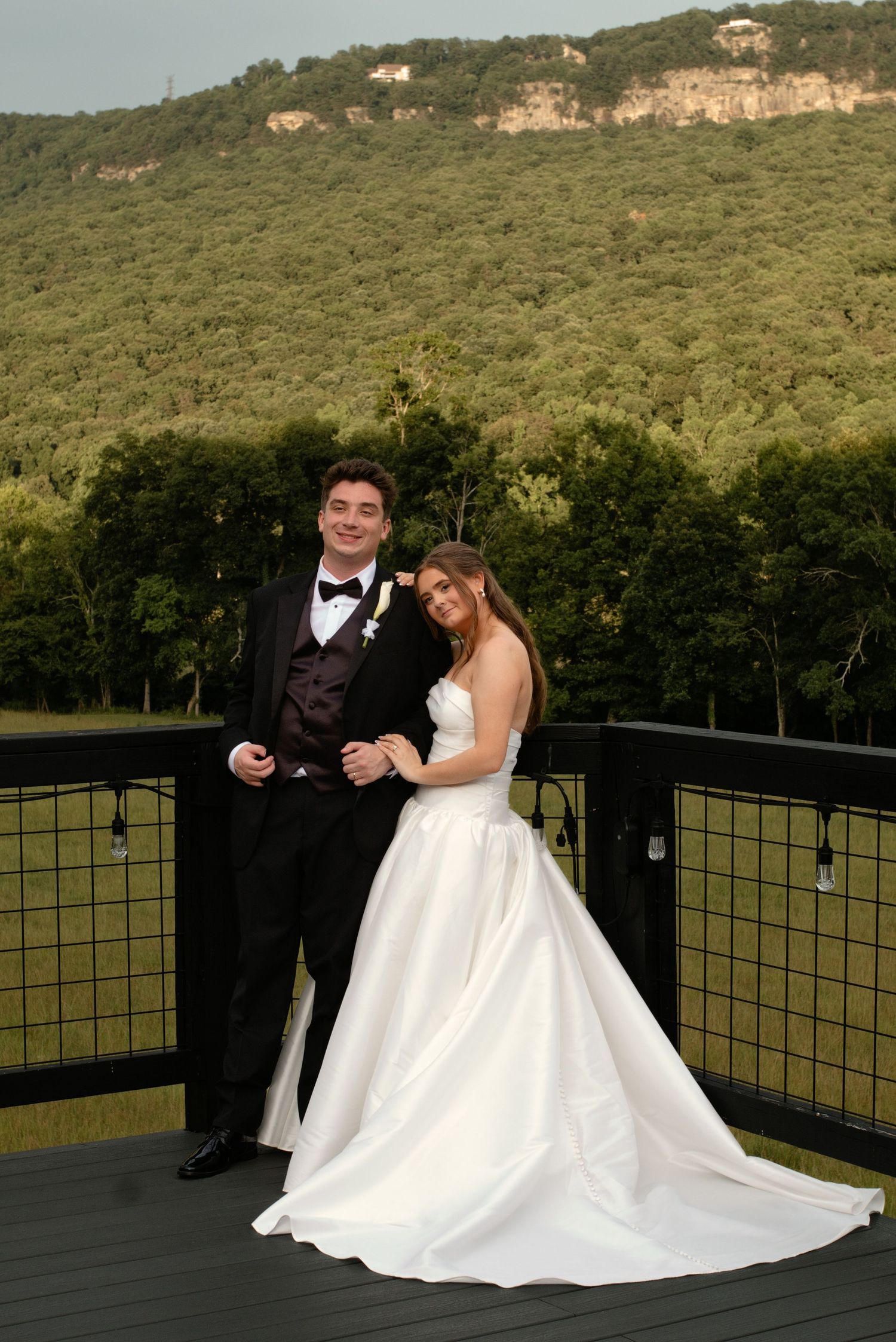 A couple in wedding attire poses on a deck with lush green mountain backdrop on their special day.