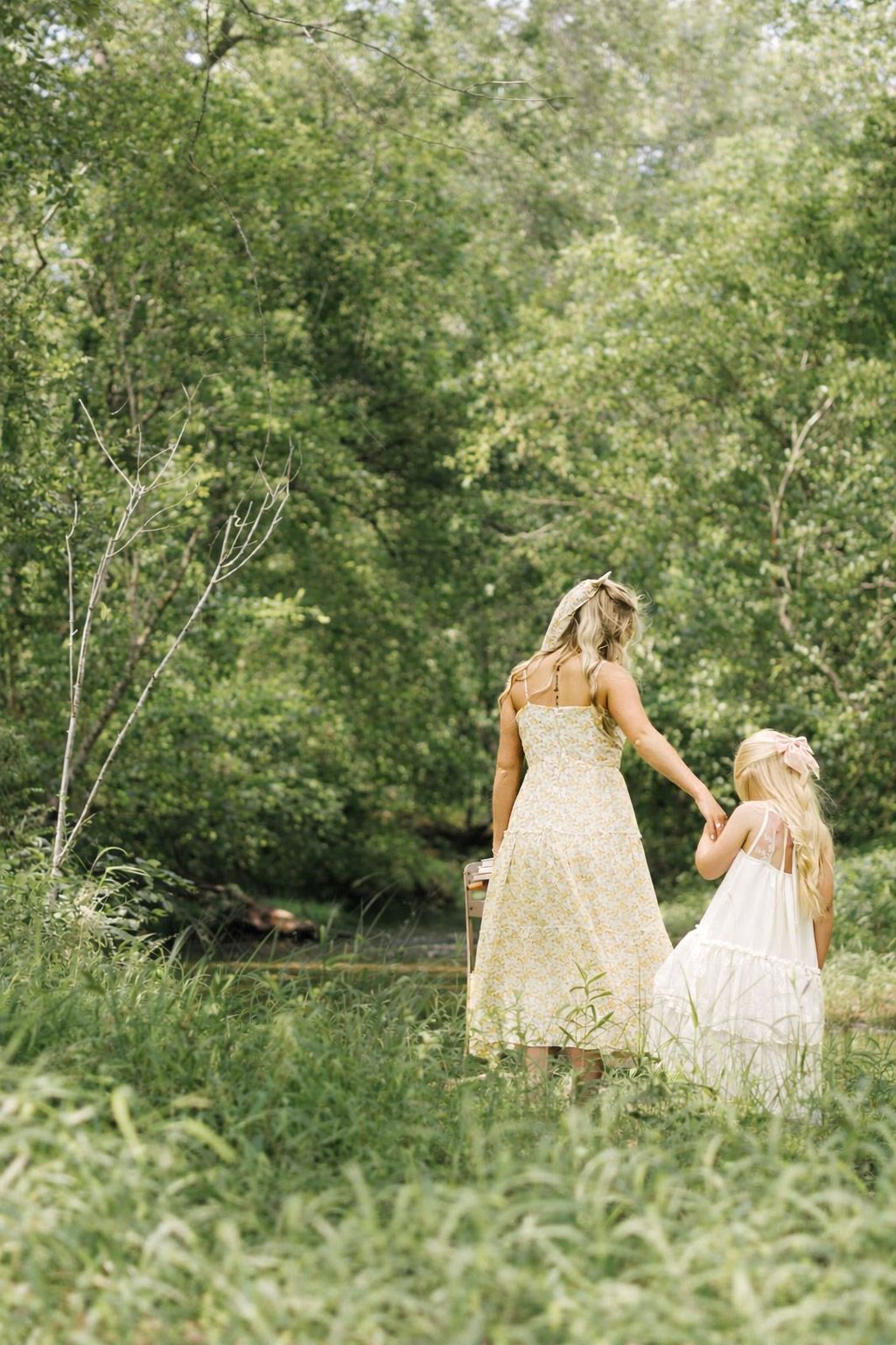 A woman and young girl in white dresses hold hands while walking through a lush green forest path.