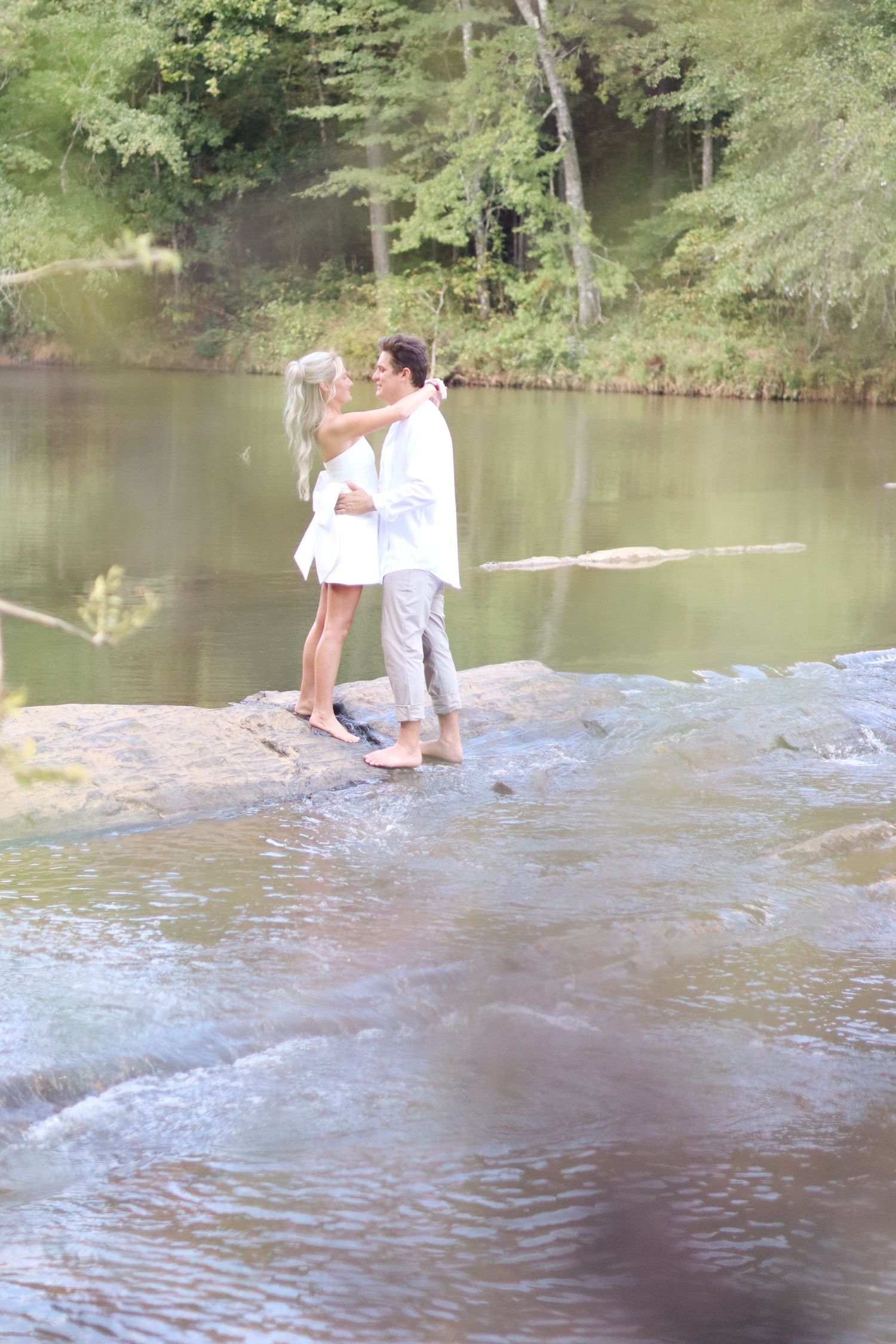 A romantic moment between a couple standing on rocks by a serene lake surrounded by trees.