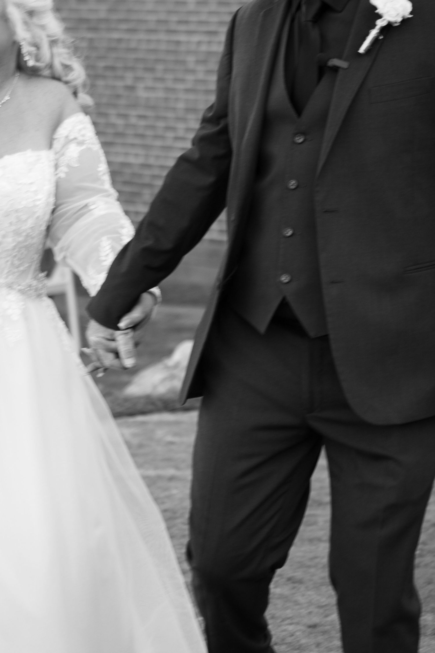 Black and white wedding photo showing bride in lace dress holding hands with groom in dark formal suit and vest.