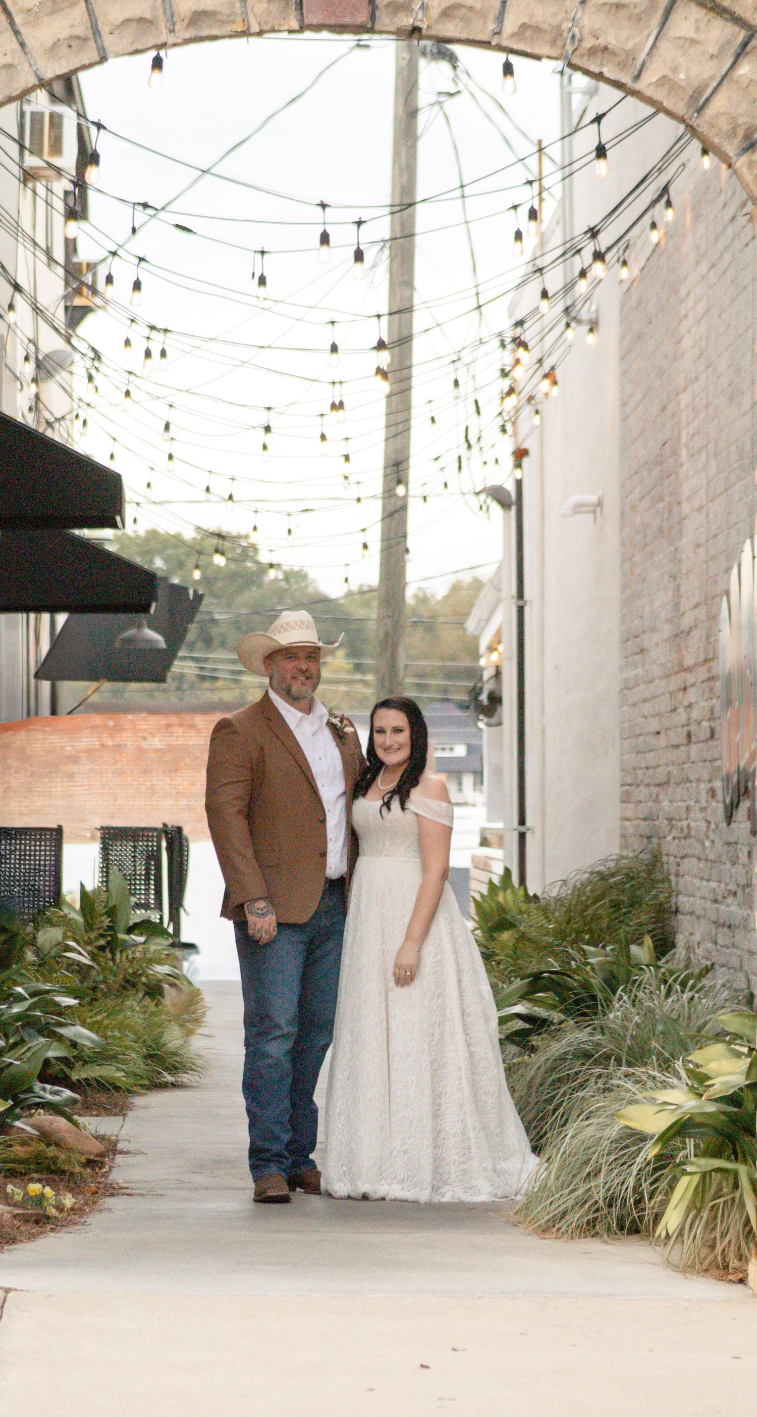 Wedding couple poses in a rustic outdoor venue with string lights and exposed brick walls.