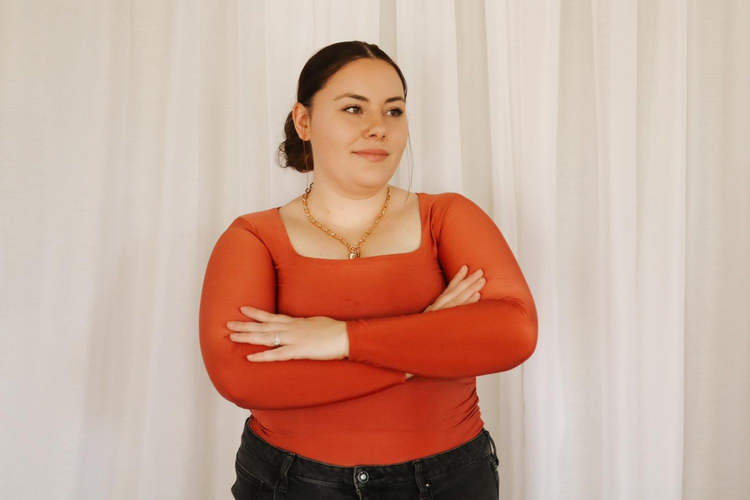 Woman in orange sweater posing with crossed arms against white background.