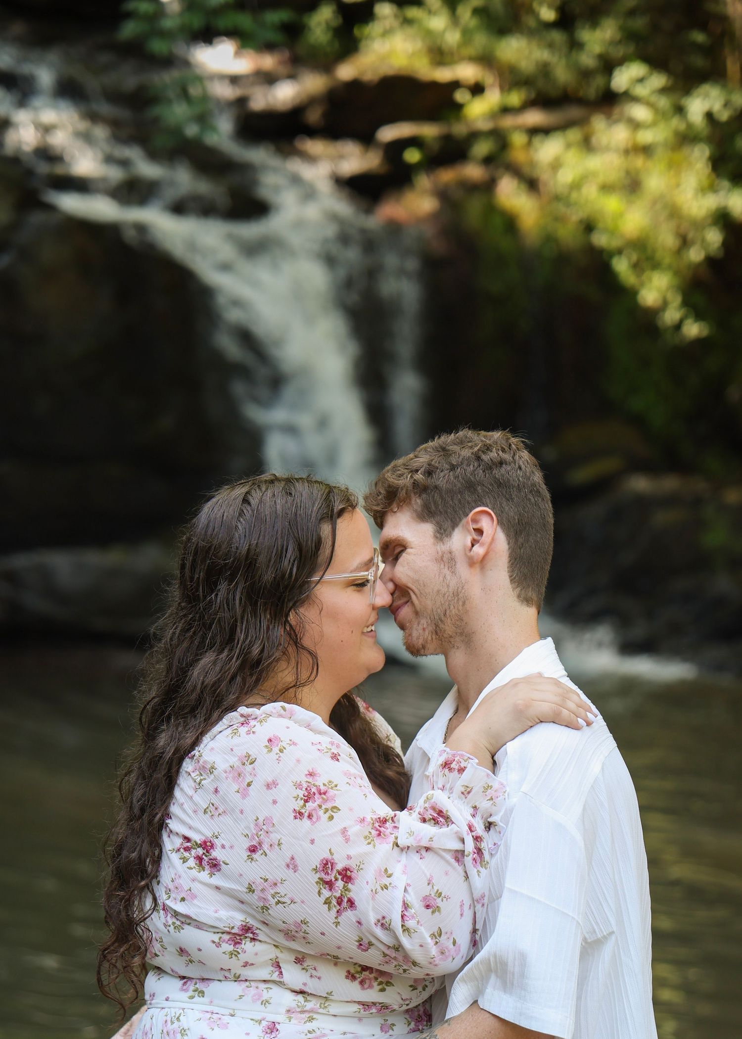 Couple embracing and smiling at each other in front of a serene waterfall, woman in floral dress, man in white shirt.