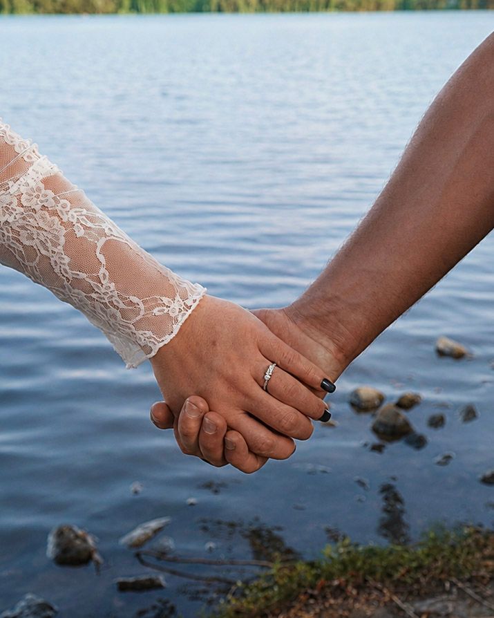 Newlywed couple holding hands by a serene lake, bride wearing lace wedding dress sleeve with diamond ring.