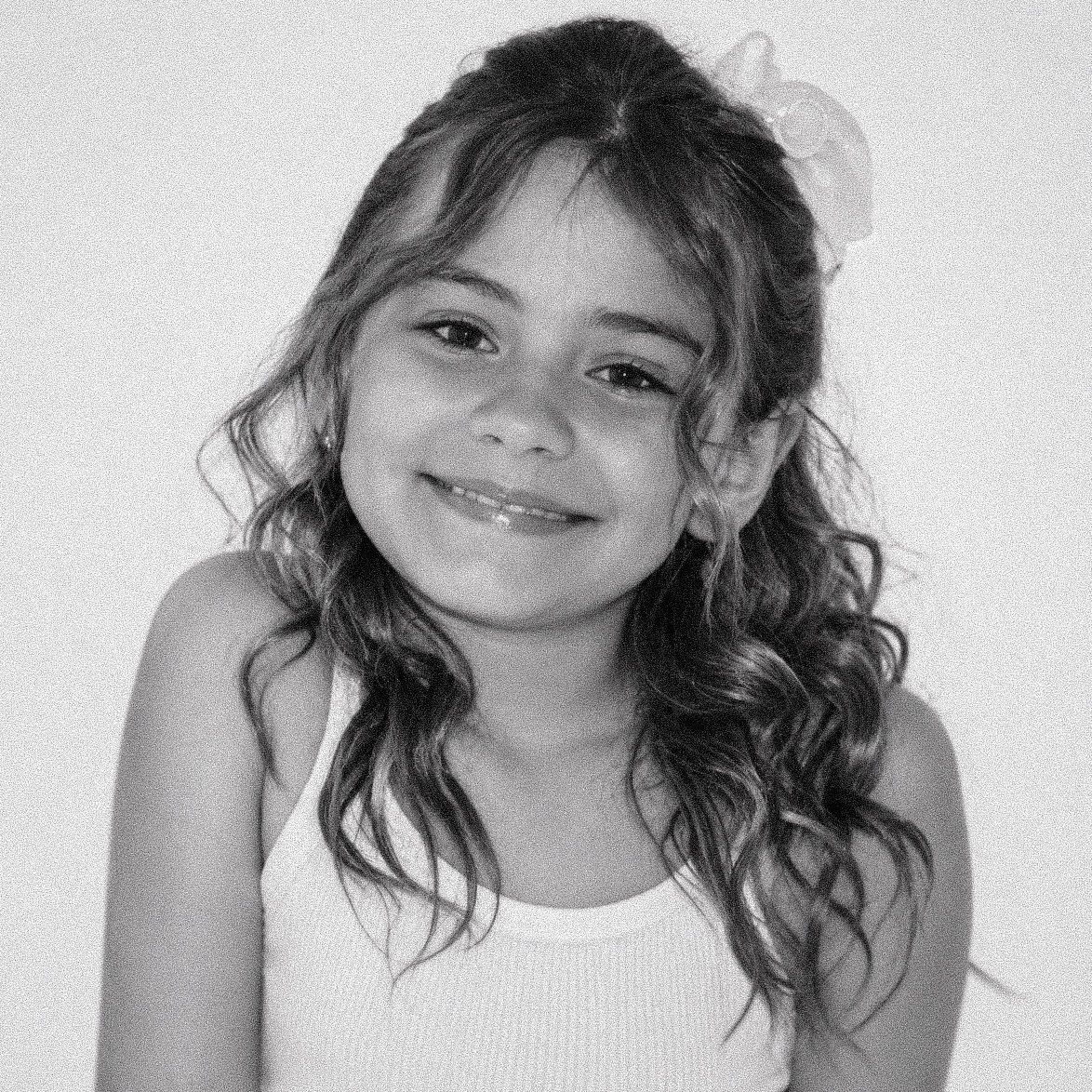 Smiling young girl with curly hair and a flower accessory poses for a black and white portrait photo.