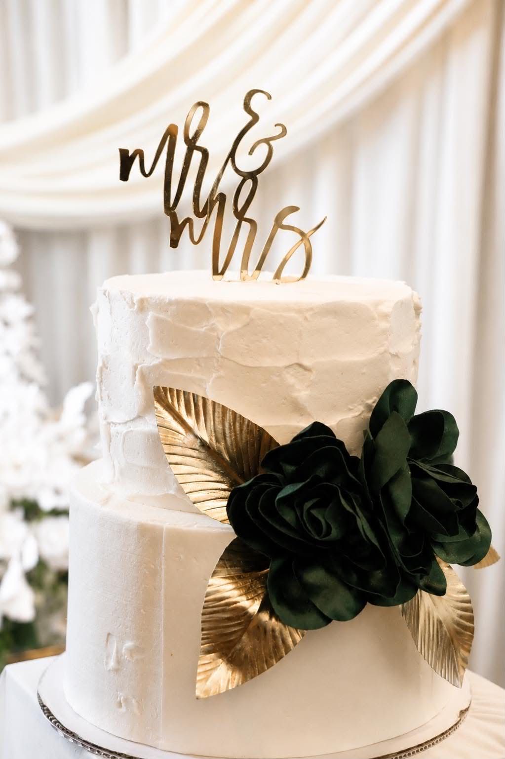 White two-tier wedding cake with gold Mr & Mrs topper, dark green floral decoration, and gold leaf accents.