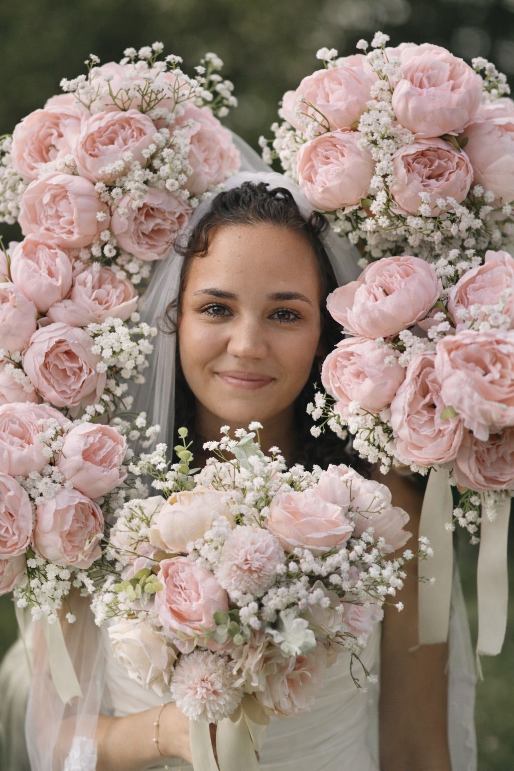 Smiling bride in white veil peers through large heart-shaped pink rose and baby's breath floral arrangement.