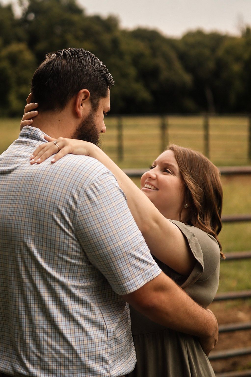 Couple embracing outdoors near a farm fence, woman smiling up at bearded man in plaid shirt, green trees in background.