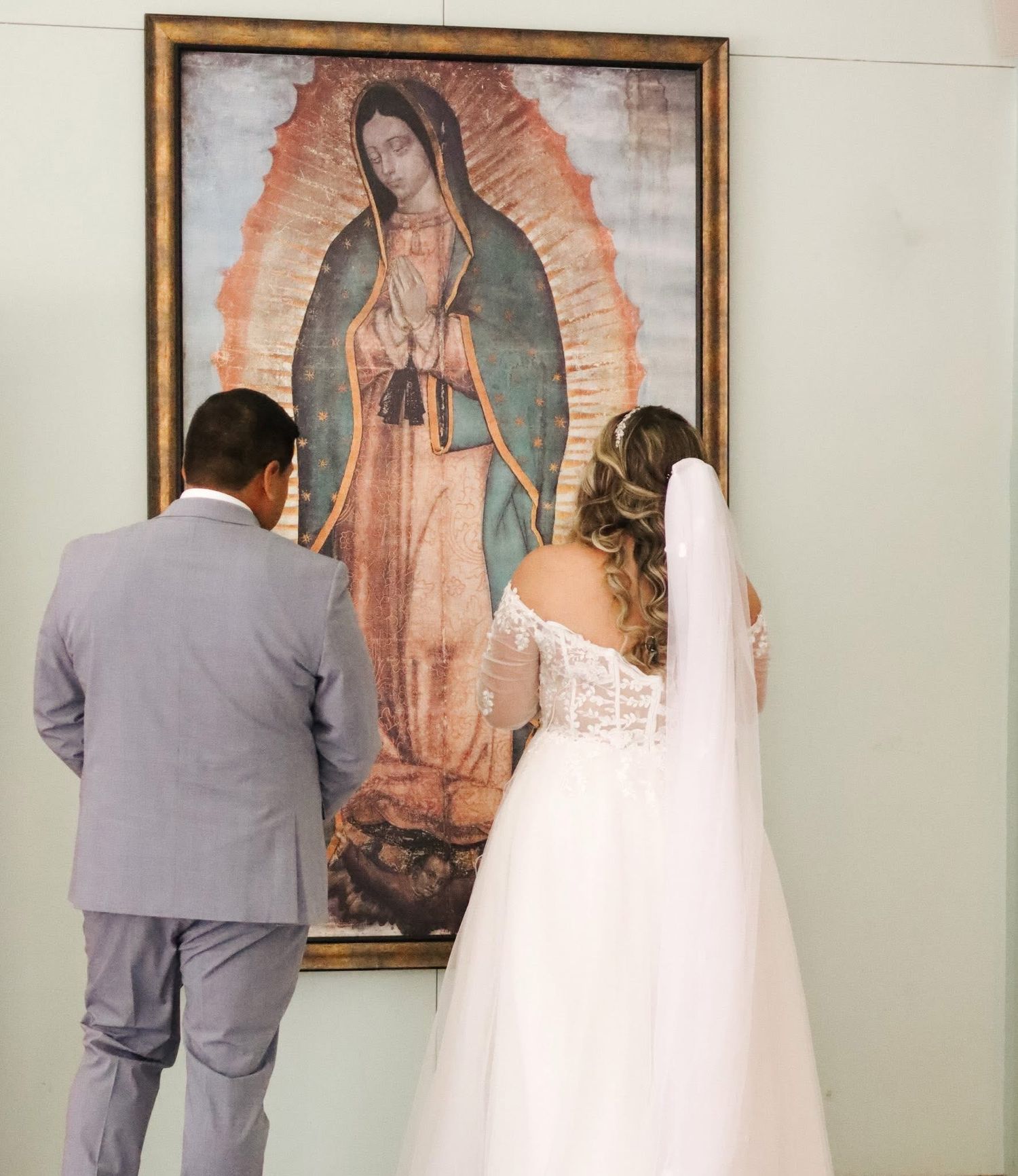 Couple praying in front of religious artwork in grey and white formal attire.