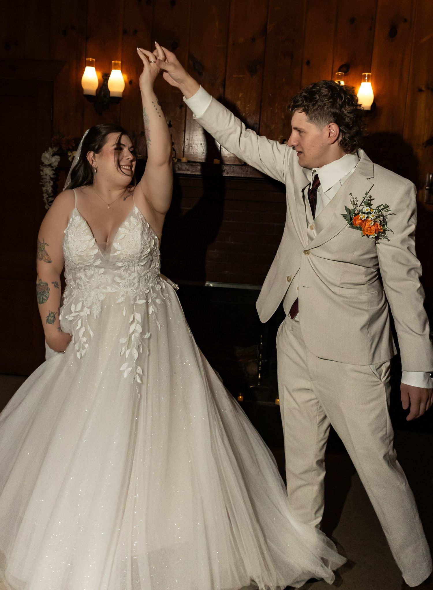 A smiling bride in a white lace gown twirls hands with a groom in a cream suit and orange boutonniere during a rustic wedding dance.