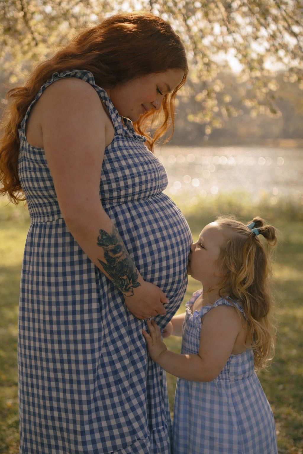 Mother and daughter in matching blue gingham dresses share a tender moment in golden sunlit field.