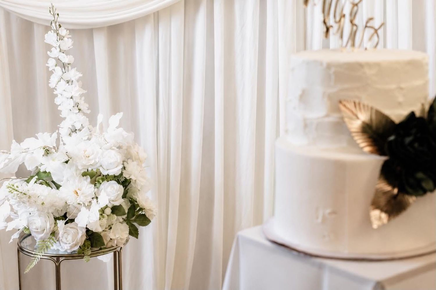 Elegant white wedding cake beside a floral arrangement of white roses and delphinium against a draped backdrop.