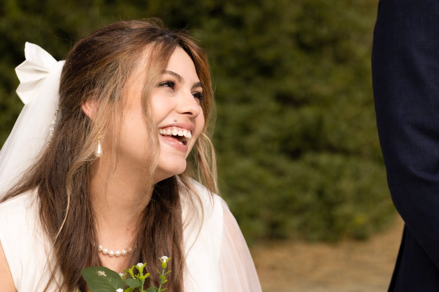 A laughing bride in a white dress poses outdoors with natural greenery in the background.
