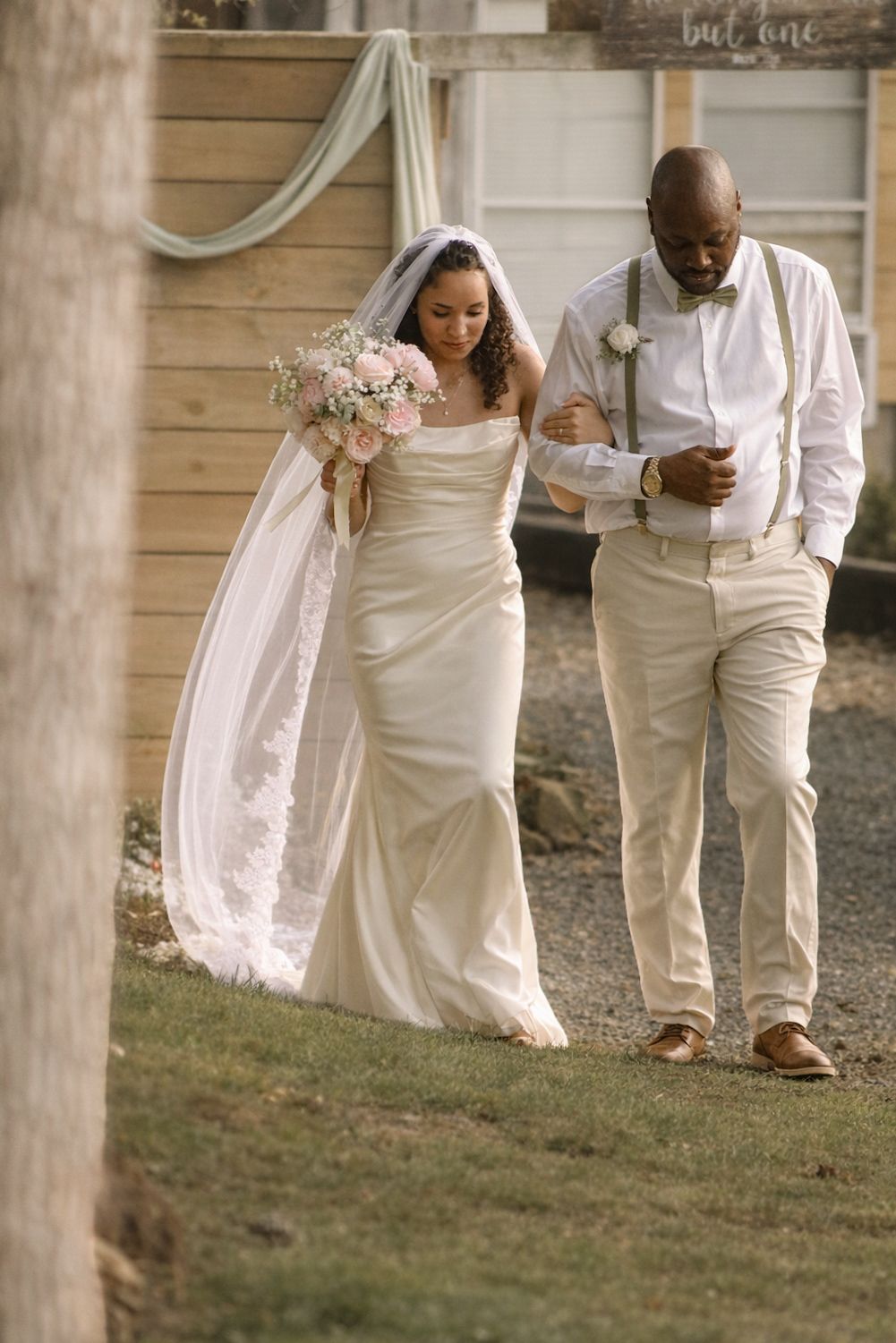 Bride in white gown and veil holding pink bouquet walks arm-in-arm with groom in khaki suspenders outdoors.