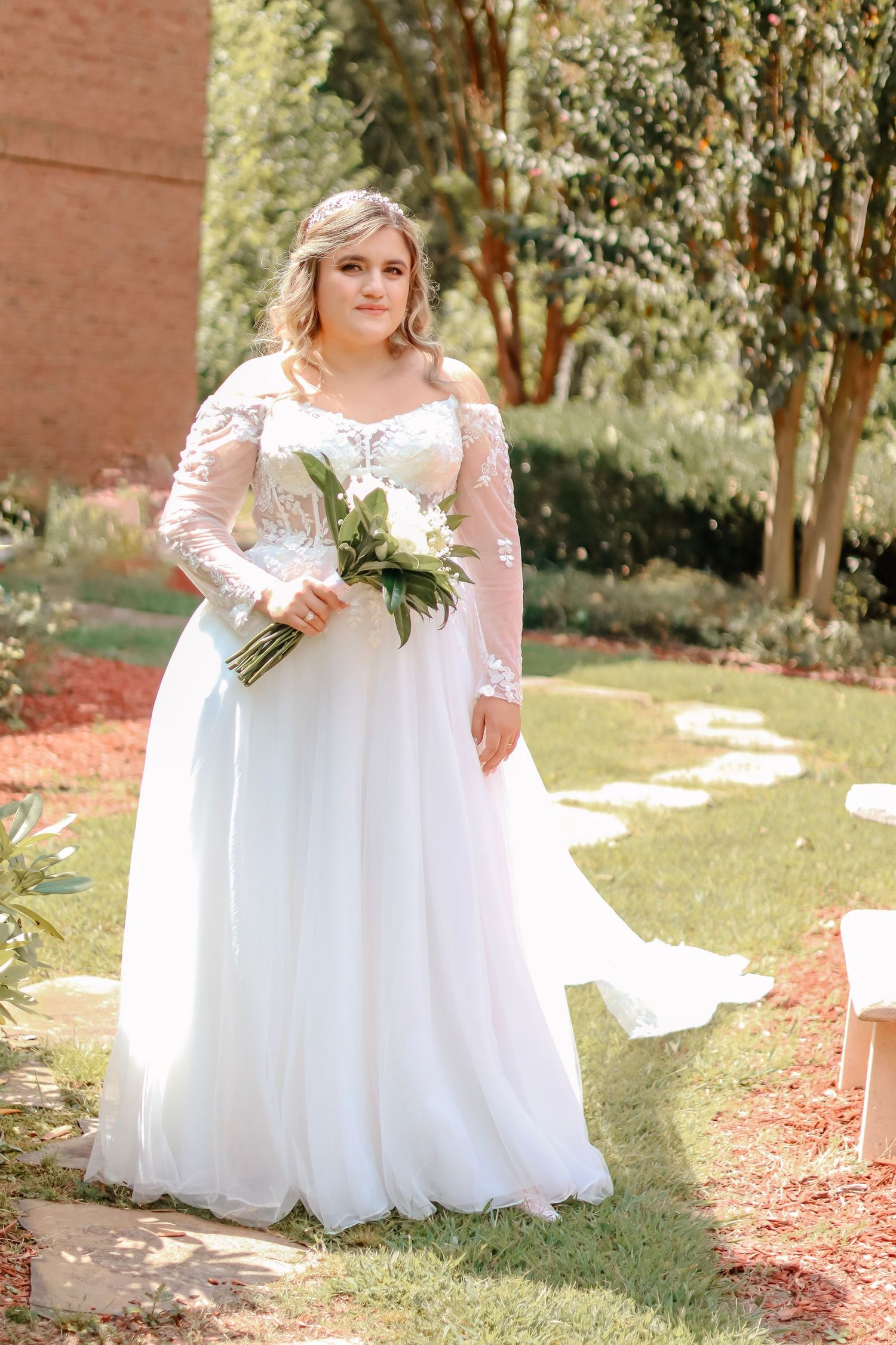Bride in lace sleeve wedding gown poses in garden setting with natural lighting.