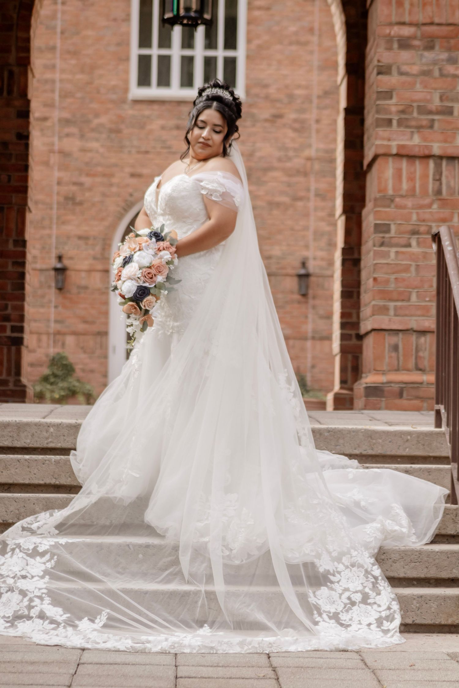 A bride in a flowing white wedding gown with lace details poses on stone steps near a brick building.