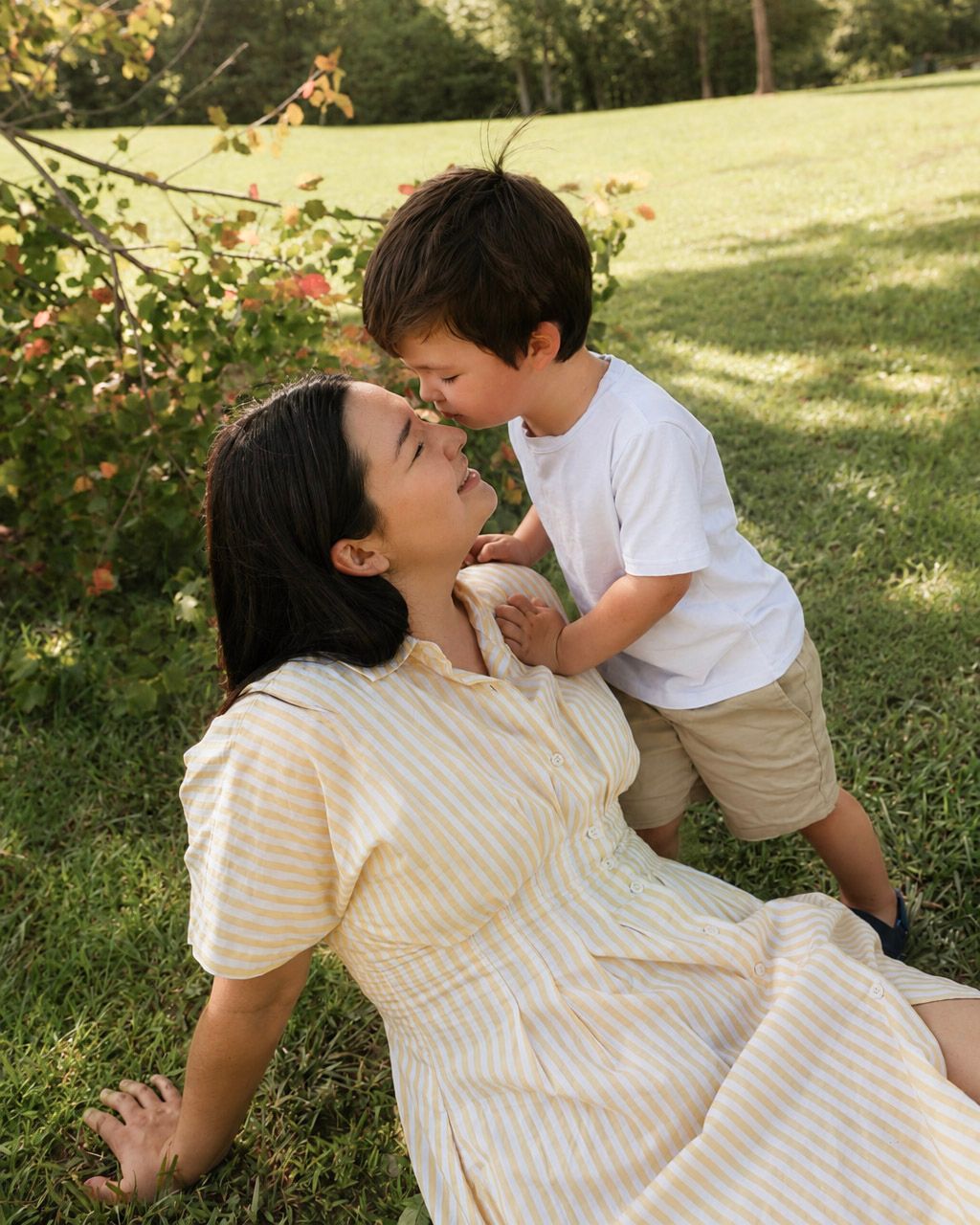A mother and young toddler boy share a sweet nose-to-nose moment on a sunny green lawn.