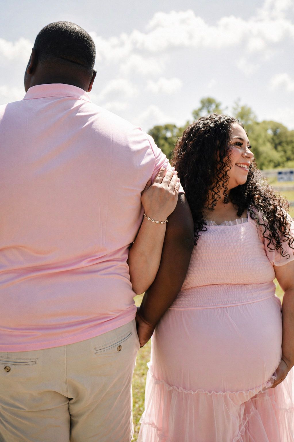 Pregnant couple in matching pink outfits standing outdoors, woman smiling with curly hair, maternity photo session.