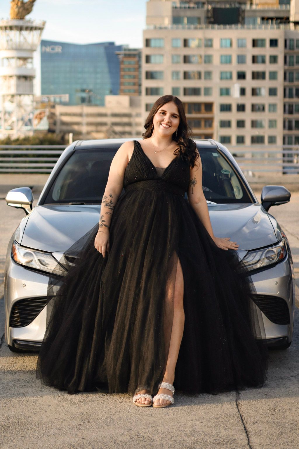 Woman in elegant black tulle gown with slit poses in front of silver car with city skyline backdrop.