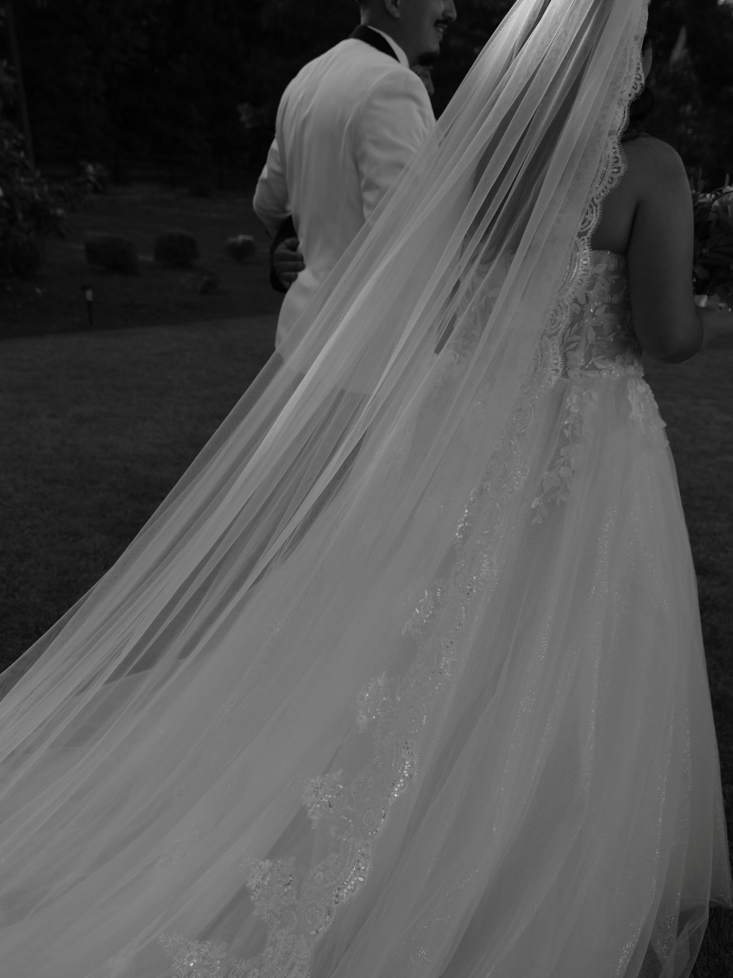 Black and white photo of a bride and groom walking, featuring an elegant lace-trimmed cathedral wedding veil.