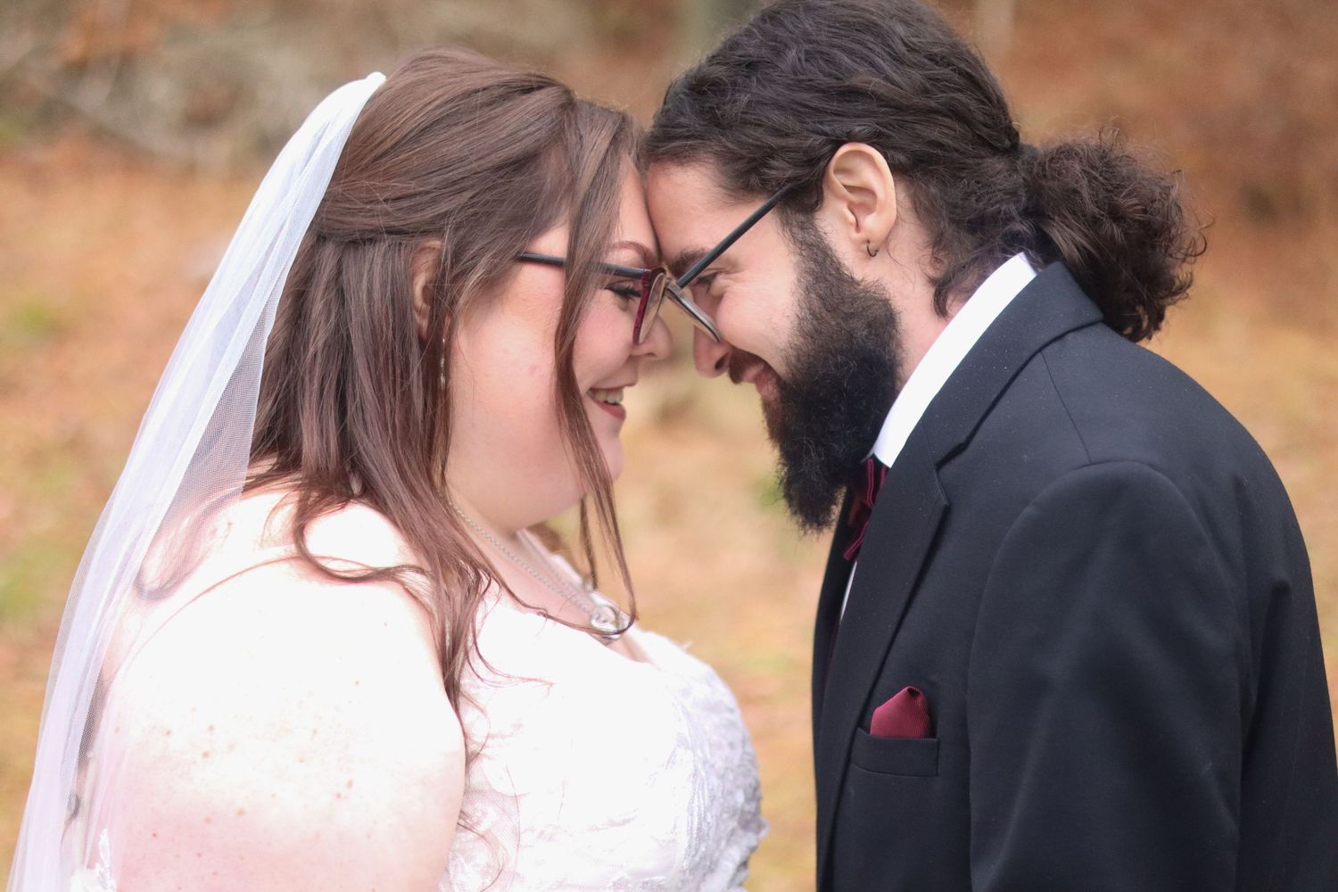 A bride in a white dress and groom in a black suit share an intimate moment outdoors with autumn colors in background.