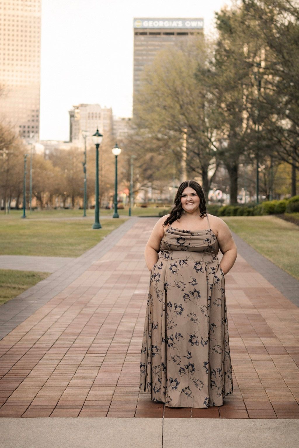 Woman in floral maxi dress posing on brick path in urban park with city buildings in background.