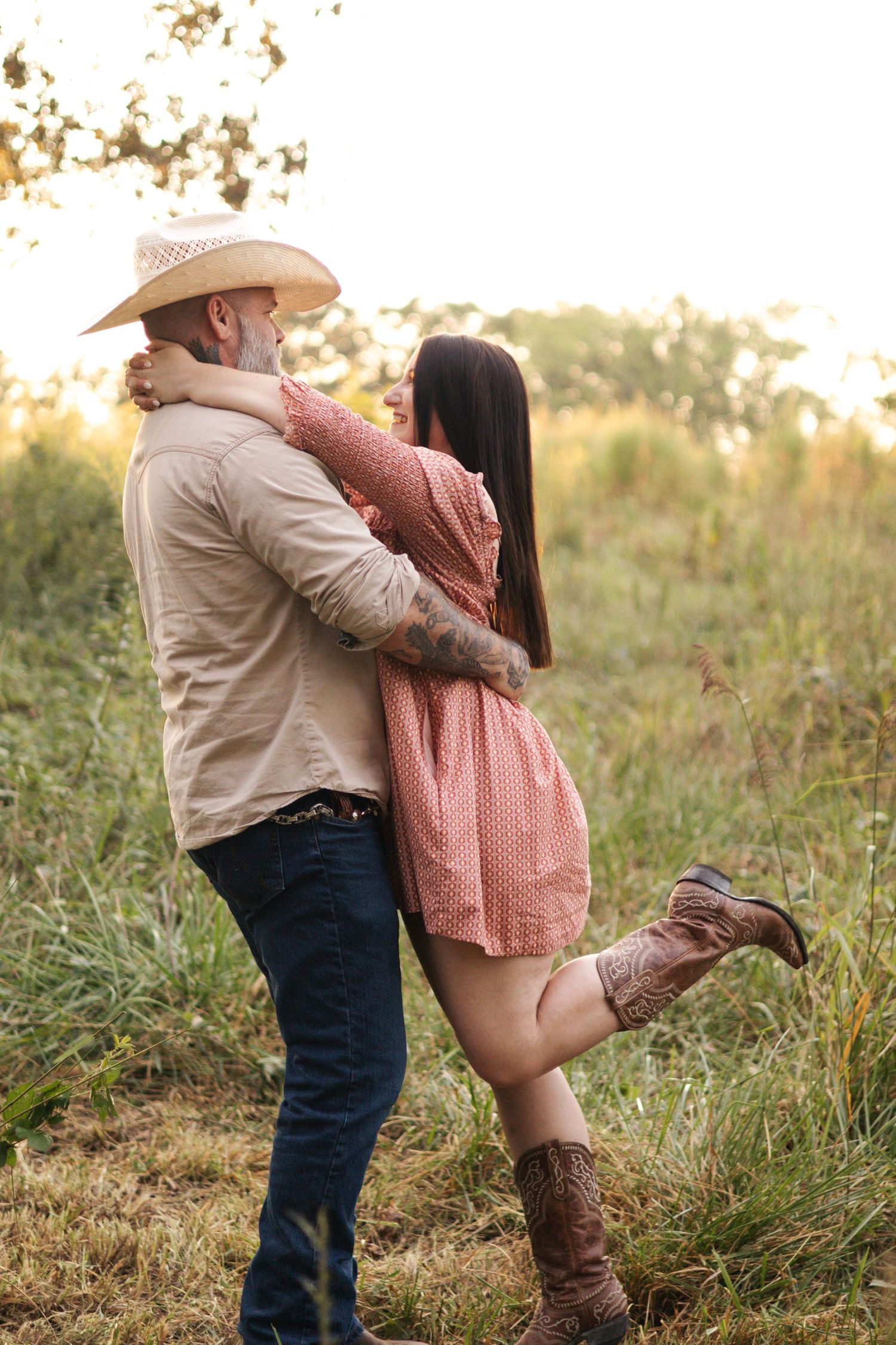 Couple in country attire embracing in a sunlit field at sunset.