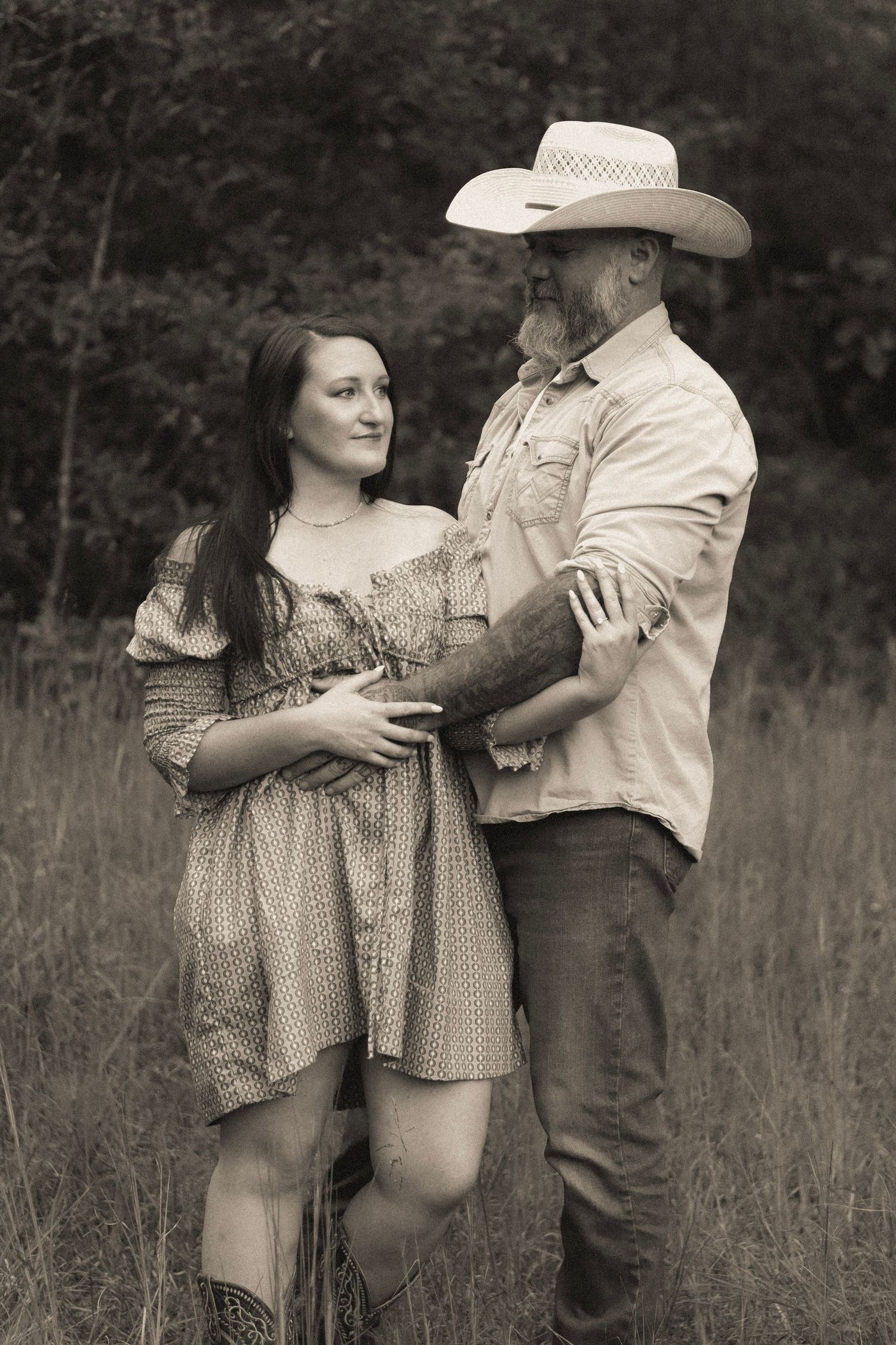 A romantic couple poses together in a field wearing country-style clothing and a cowboy hat.