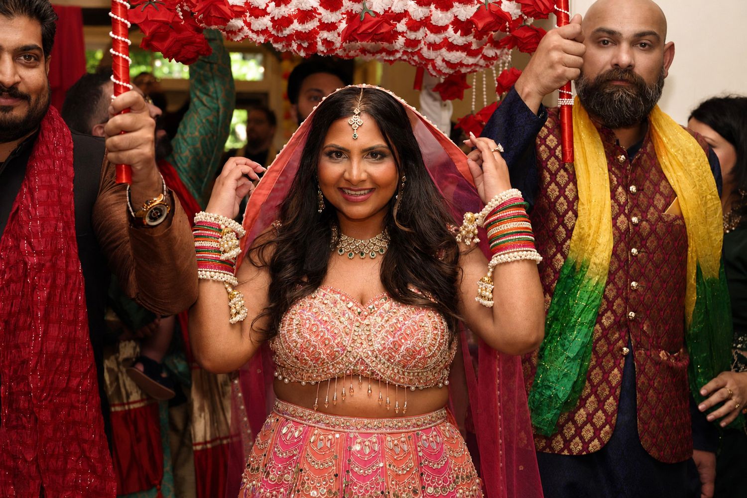 Bride in pink lehenga walks under floral canopy during Indian wedding ceremony.