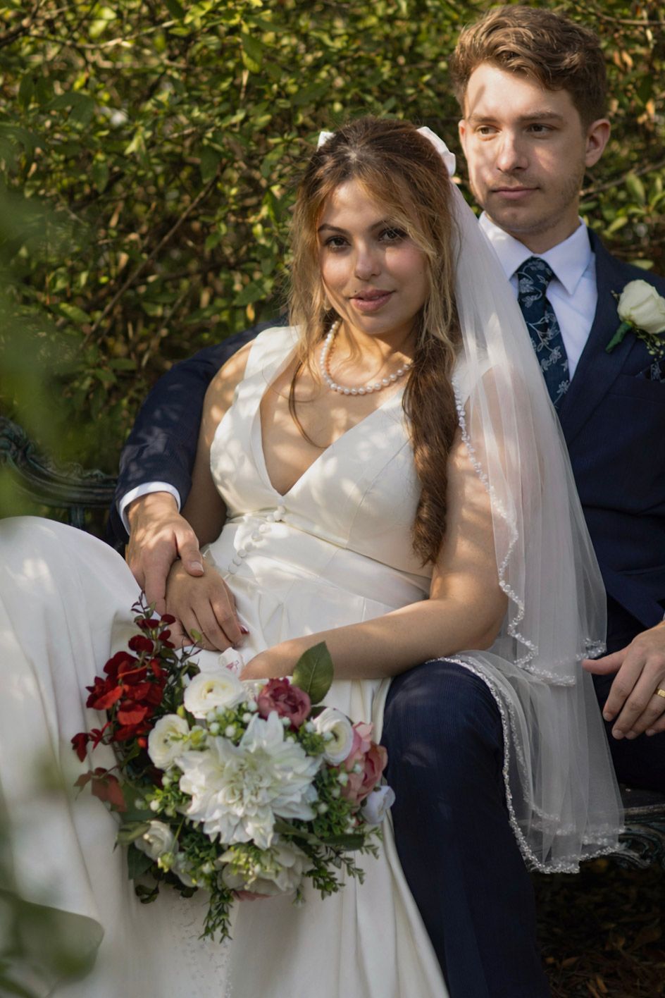 A bride in a white dress and groom in navy suit pose together outdoors with red and white floral bouquet.
