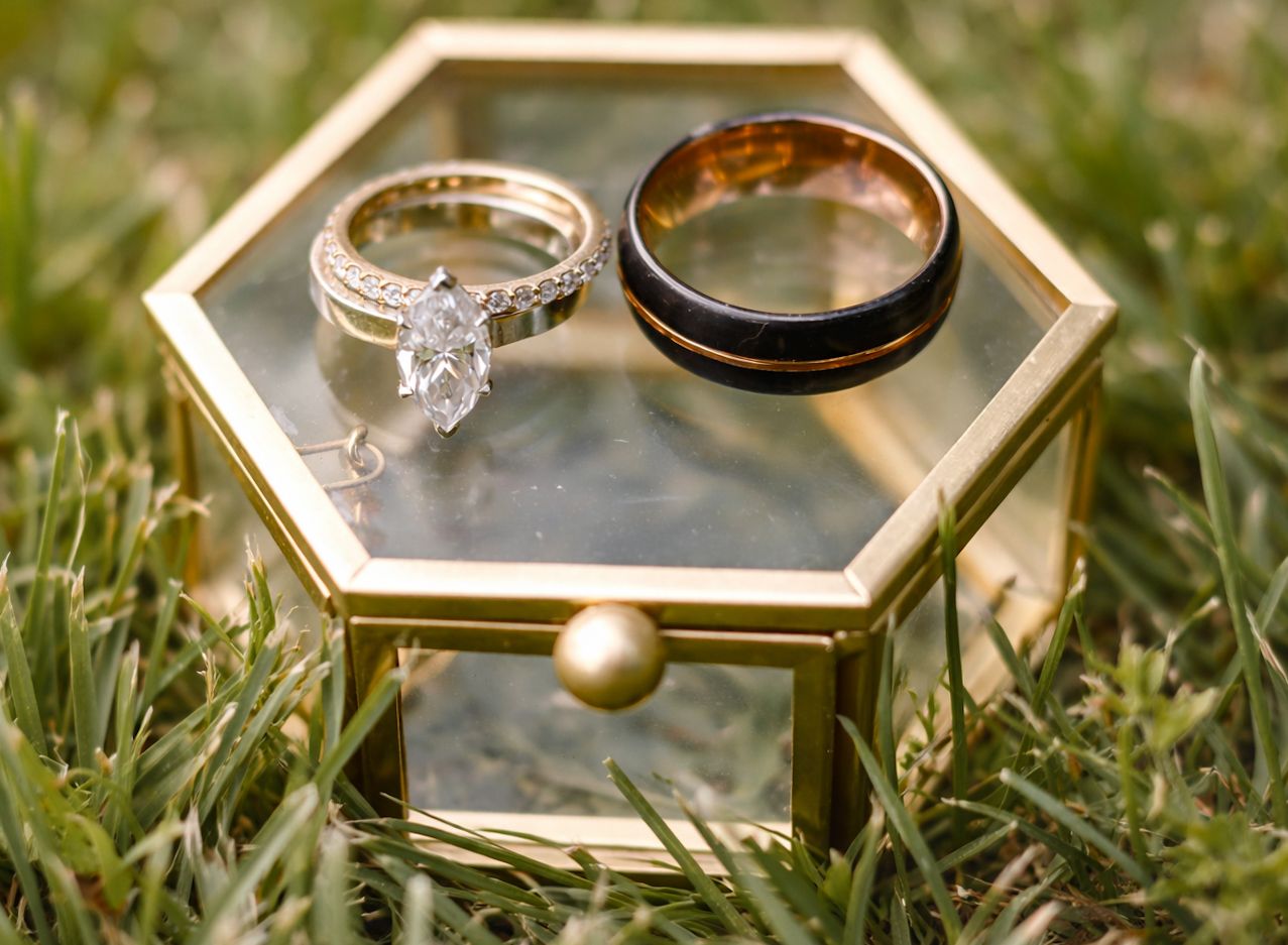 Wedding rings displayed in hexagonal gold glass box on grass, featuring diamond engagement ring and black band.