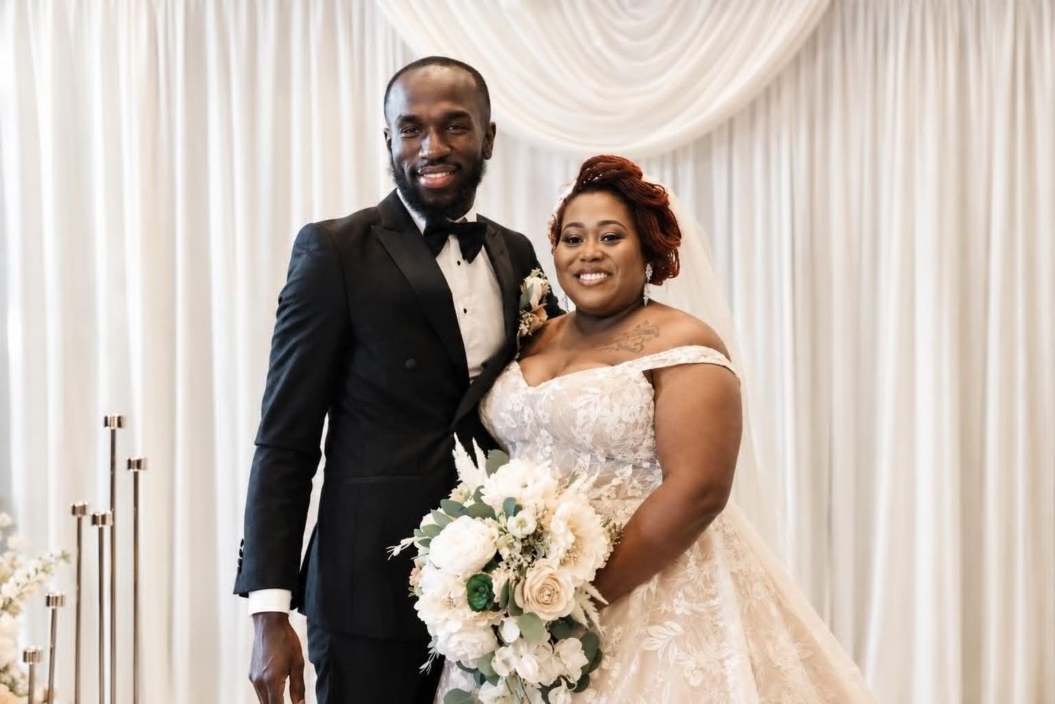 Smiling bride and groom pose together at elegant wedding ceremony, bride holding white floral bouquet in beaded gown.