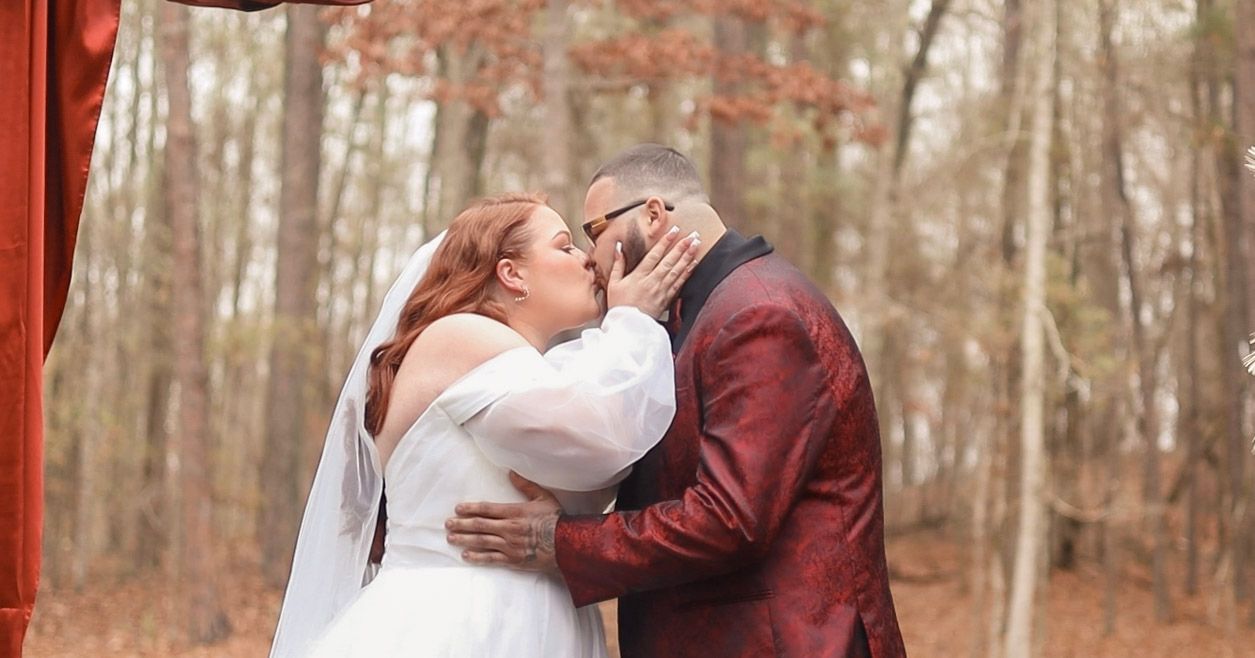 A couple in wedding attire embraces in a scenic autumn forest setting with warm orange foliage in the background.