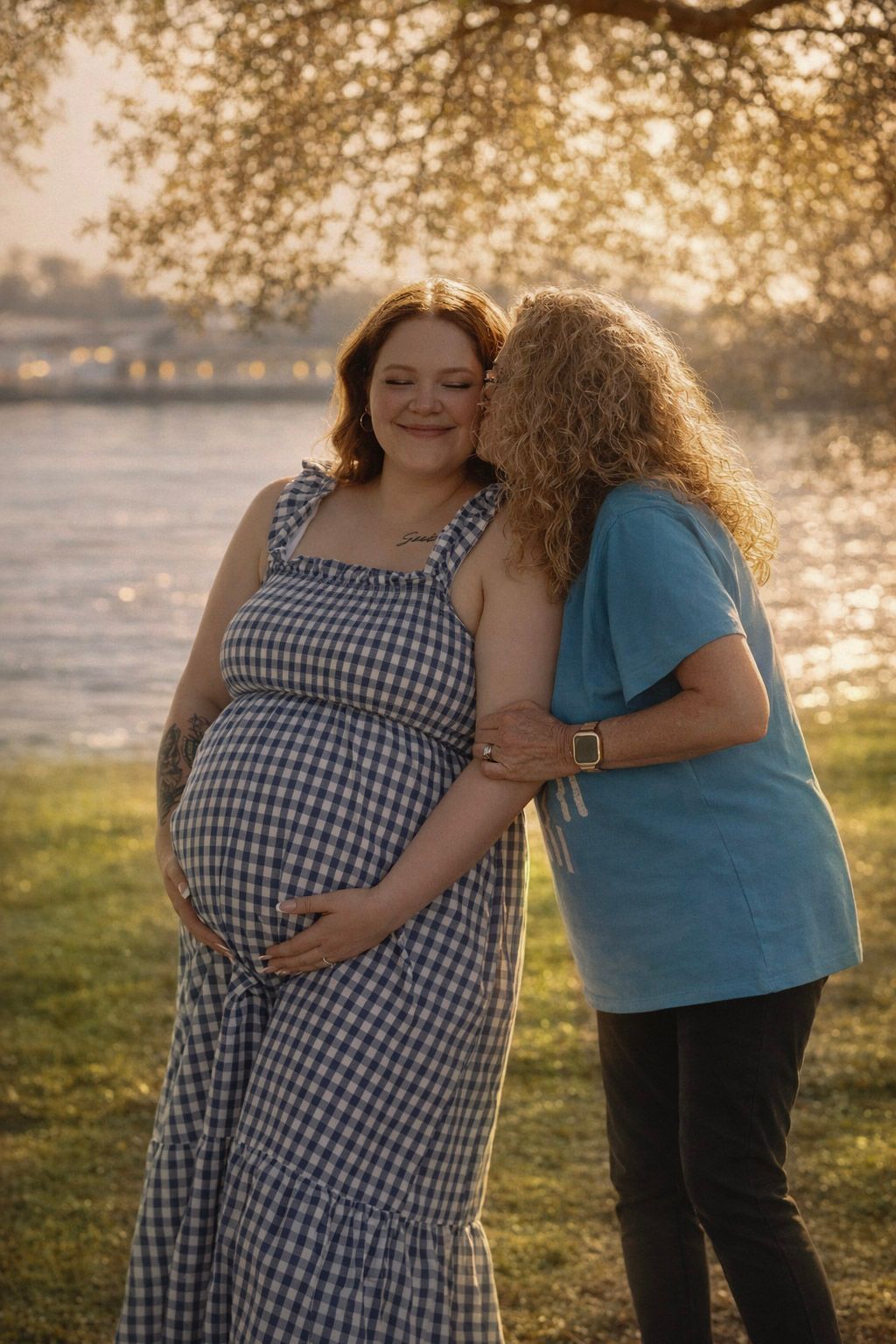 A pregnant woman in a gingham dress cradles her belly while another woman kisses her cheek outdoors at golden hour.