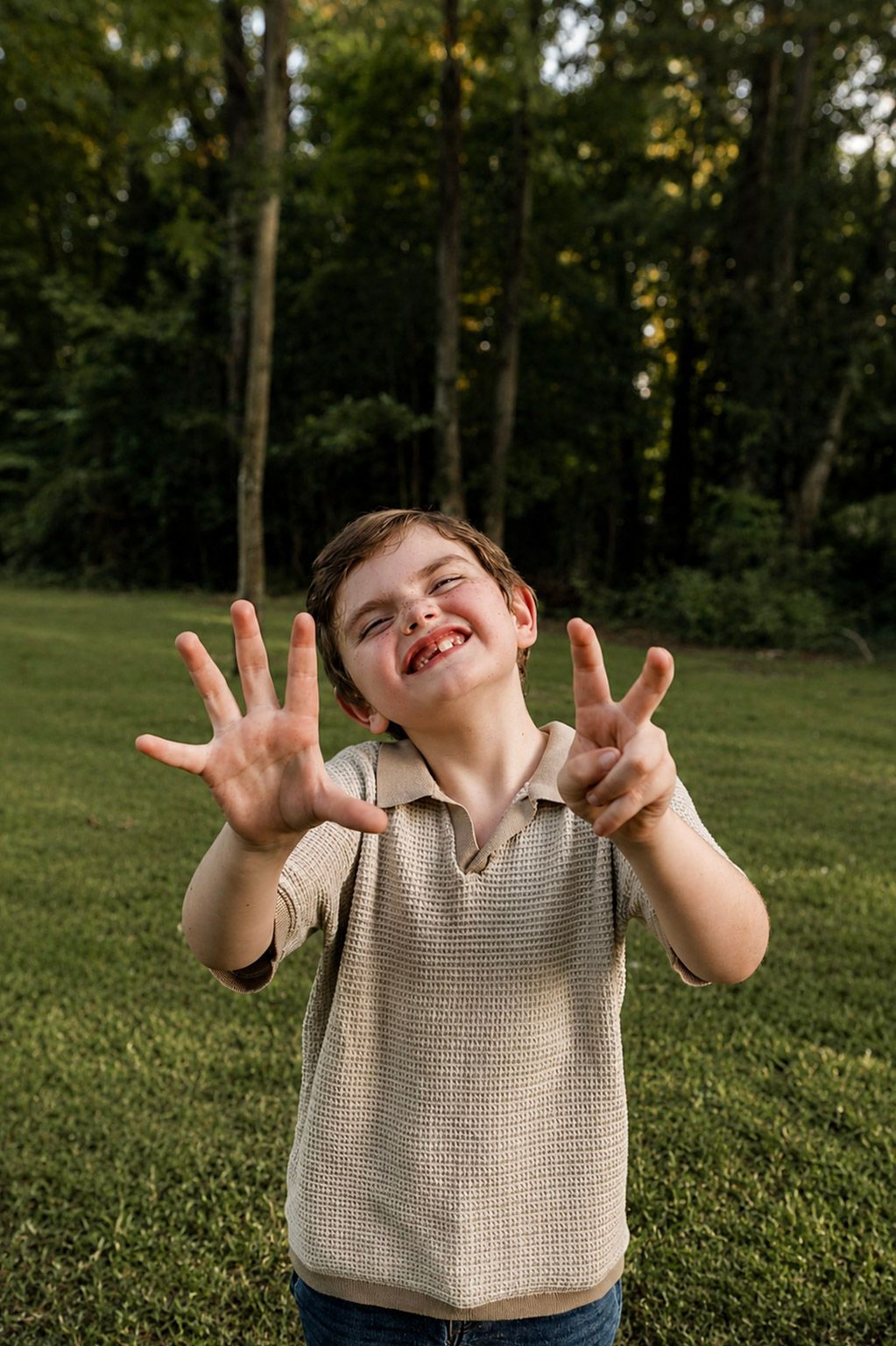 A young boy with a missing tooth grins outdoors, holding up seven fingers in a grassy backyard surrounded by trees.
