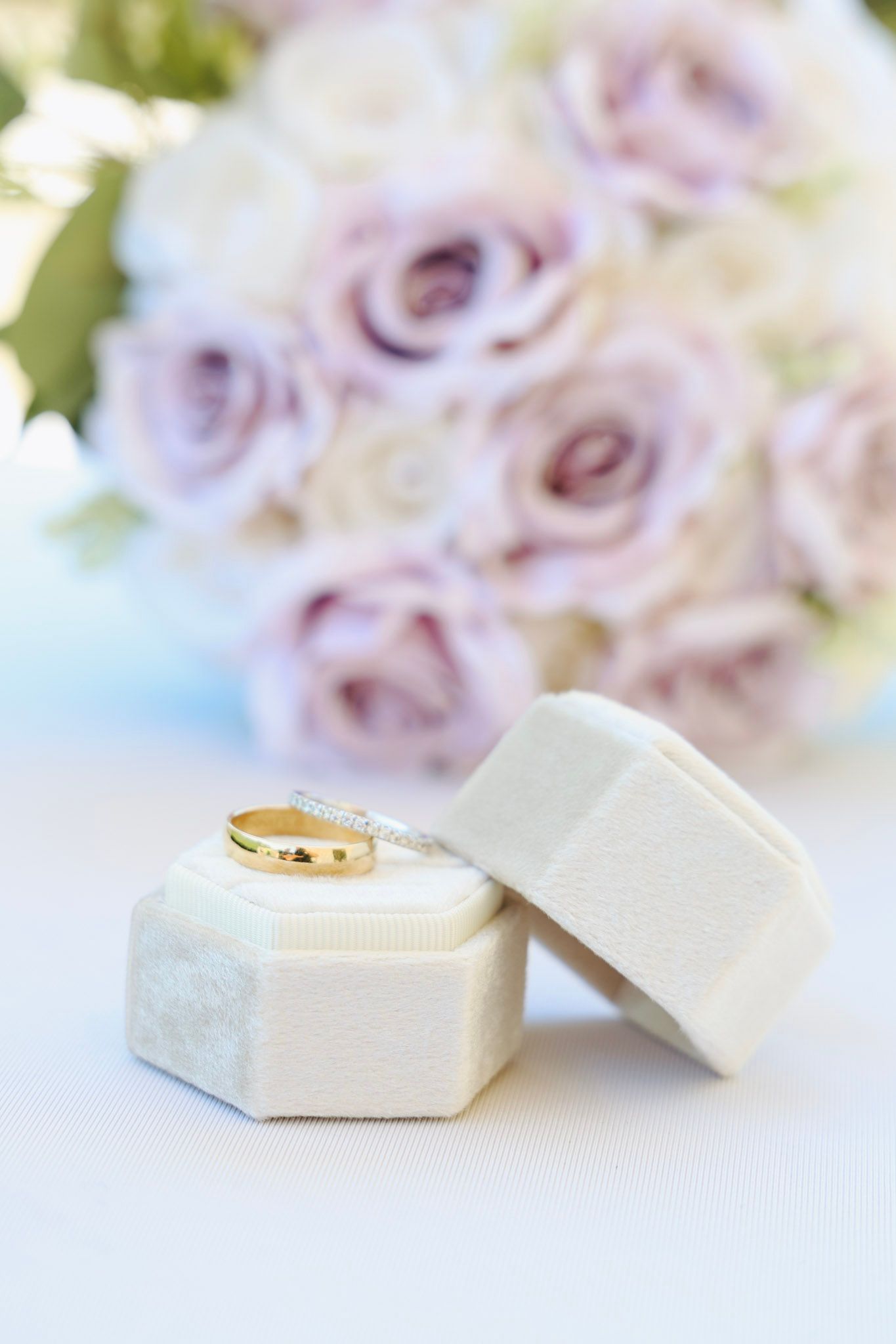 Gold and diamond wedding rings resting on a white velvet hexagonal ring box with blush roses bouquet in background.