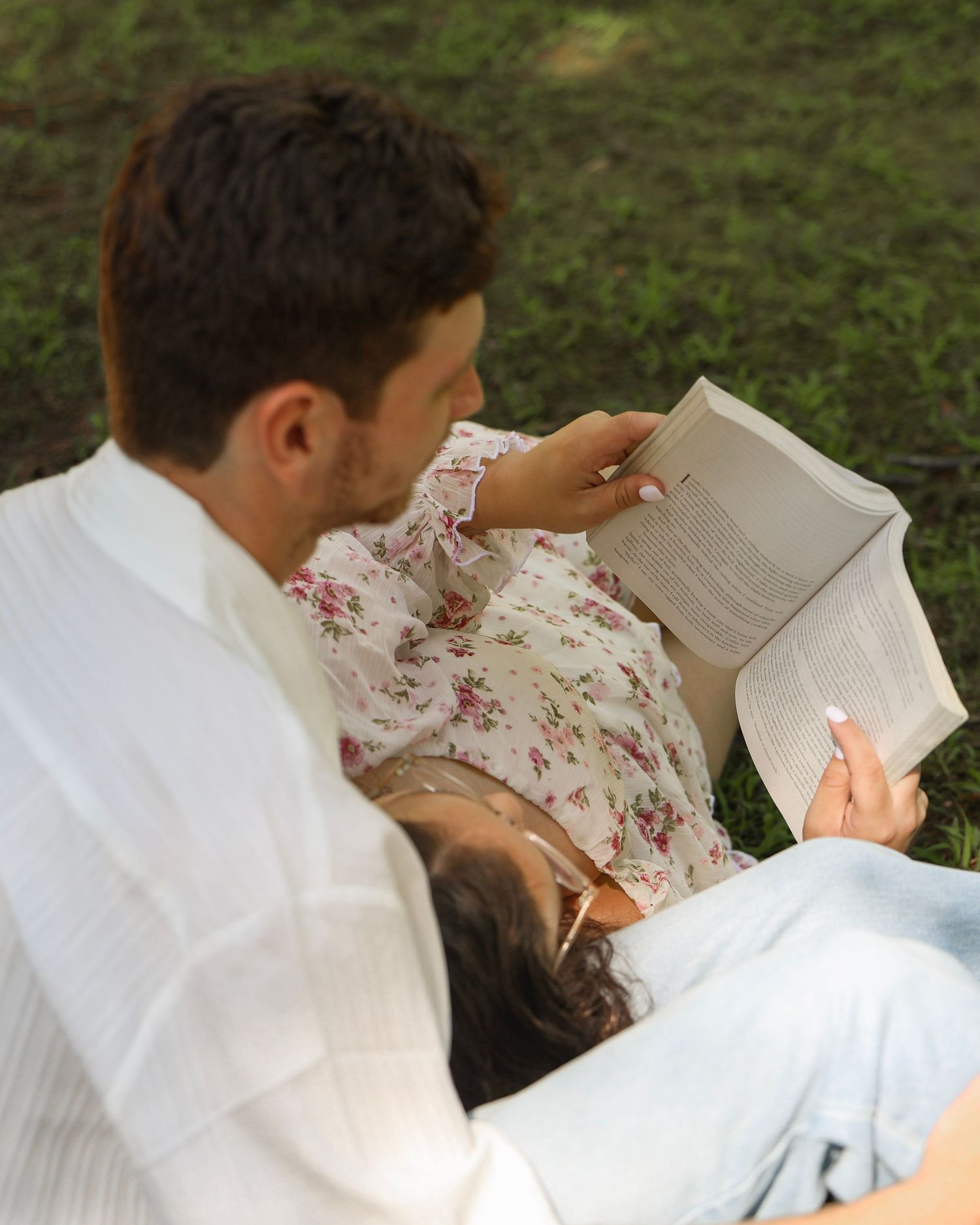 Couple relaxing outdoors on grass, woman in floral dress reading a book while resting on man's lap.