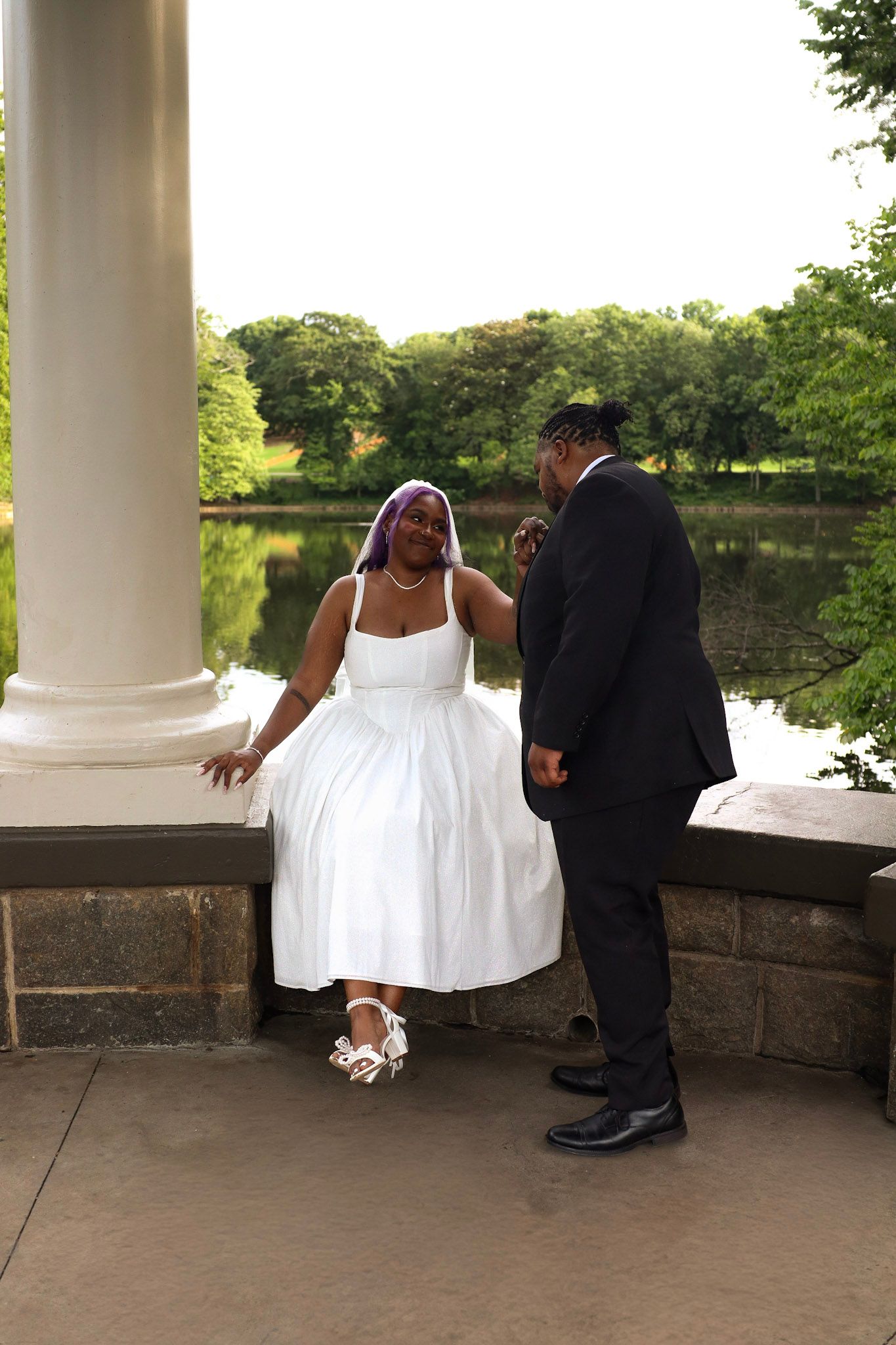 Bride and groom pose at an outdoor pavilion near a pond, bride in white midi dress and veil, groom in black suit.