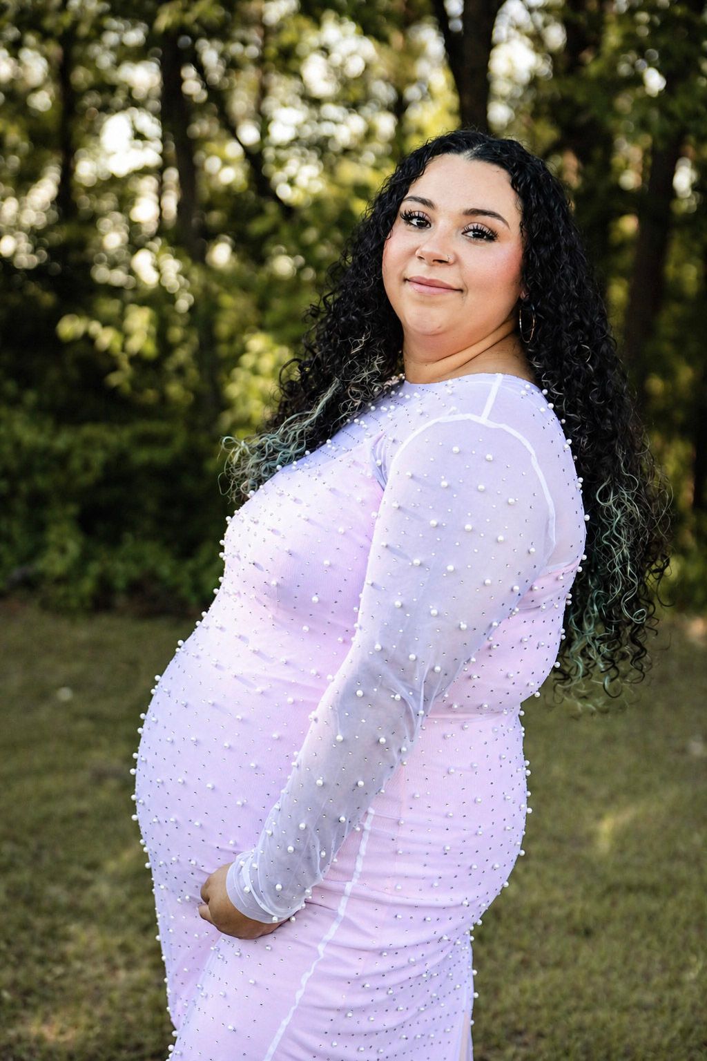 Pregnant woman in pearl-embellished white mesh dress posing outdoors with curly black hair and green trees behind her.