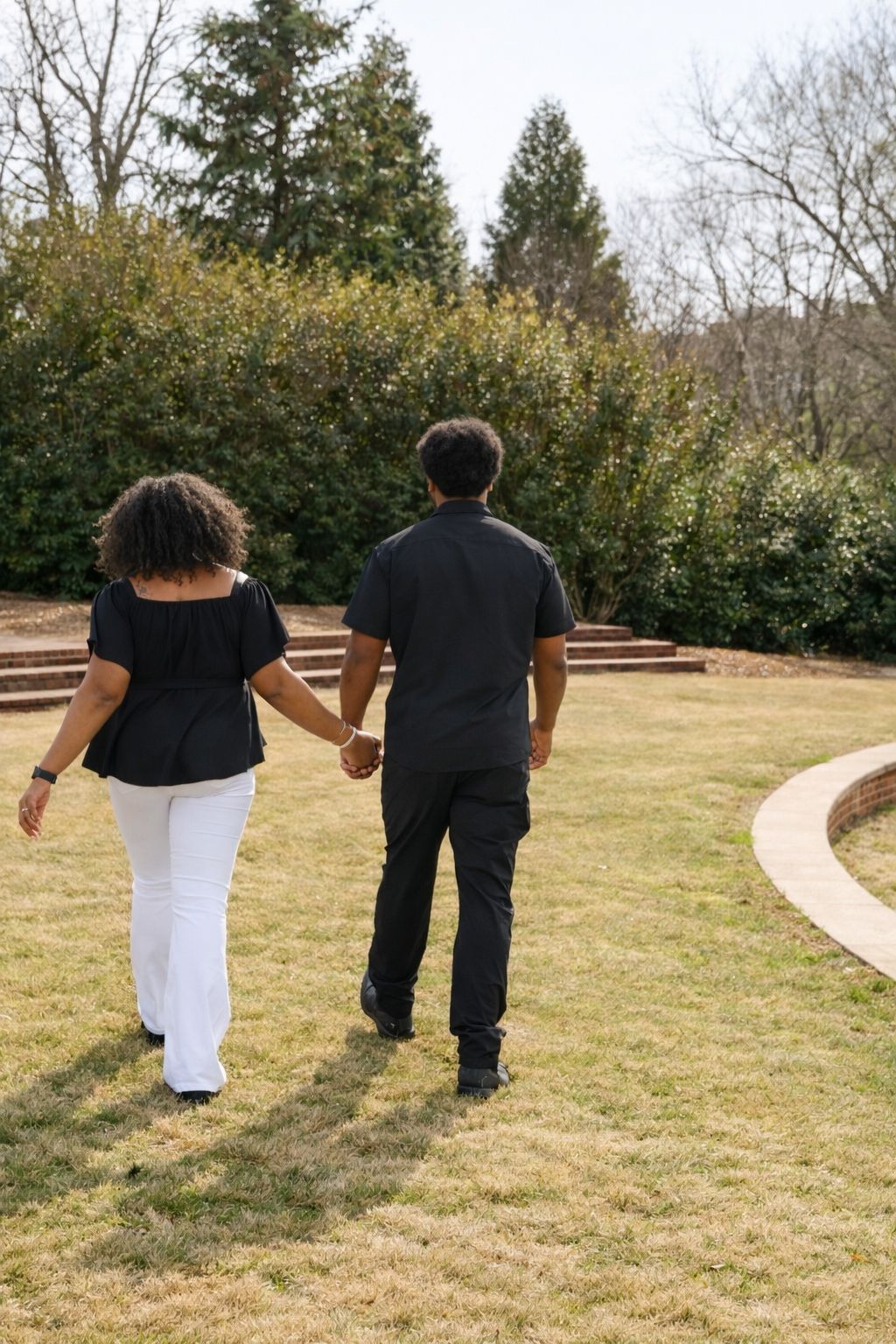 A couple walks hand in hand along a winding path through a grassy field on a sunny day.