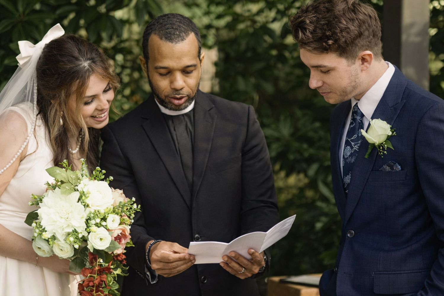 Wedding party members in formal attire read from papers during an outdoor ceremony in a garden setting.