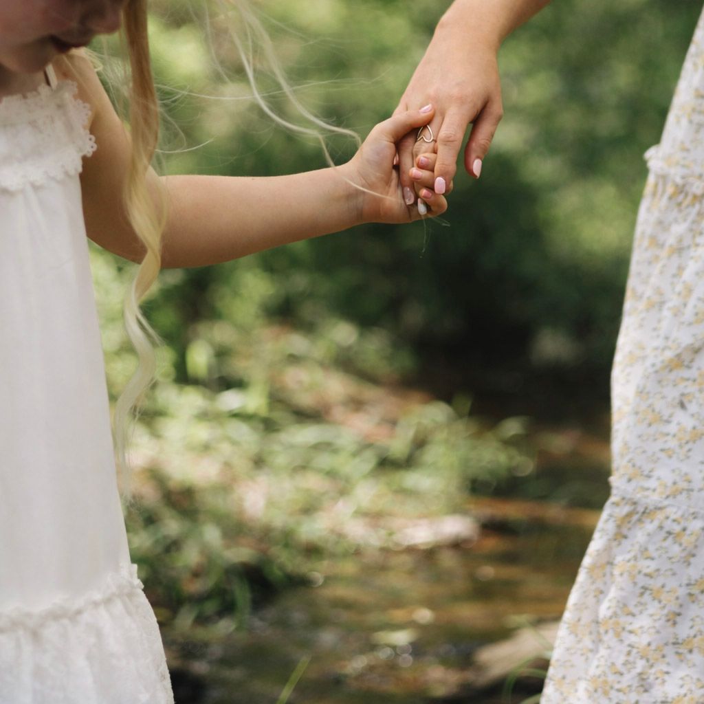 A child and adult holding hands outdoors near a stream, both wearing white dresses, surrounded by lush green foliage.
