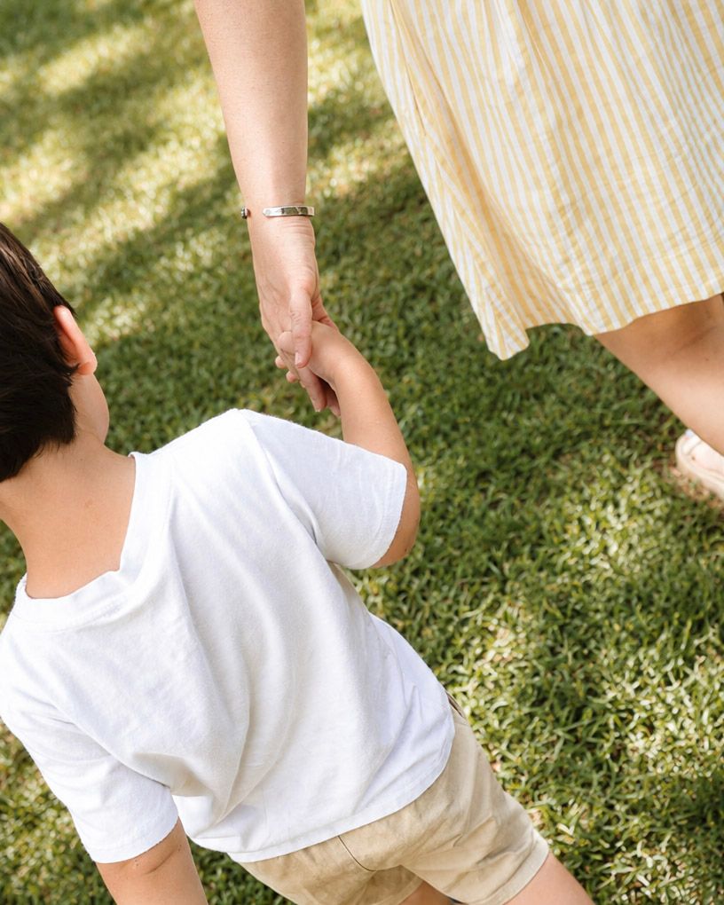 A child in a white shirt holds hands with an adult in a yellow striped dress on green grass.