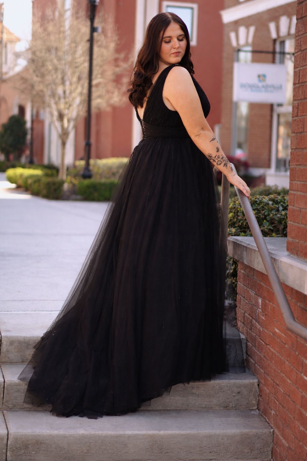 A flowing black tulle formal gown is showcased against a brick building backdrop in an outdoor photoshoot.