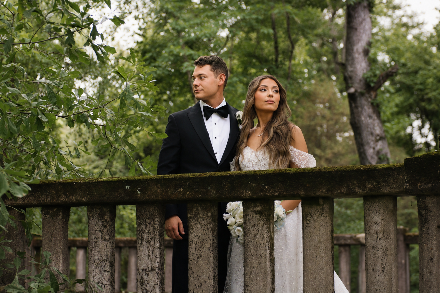 Bride and groom in formal wedding attire standing on a moss-covered stone bridge surrounded by lush green trees.