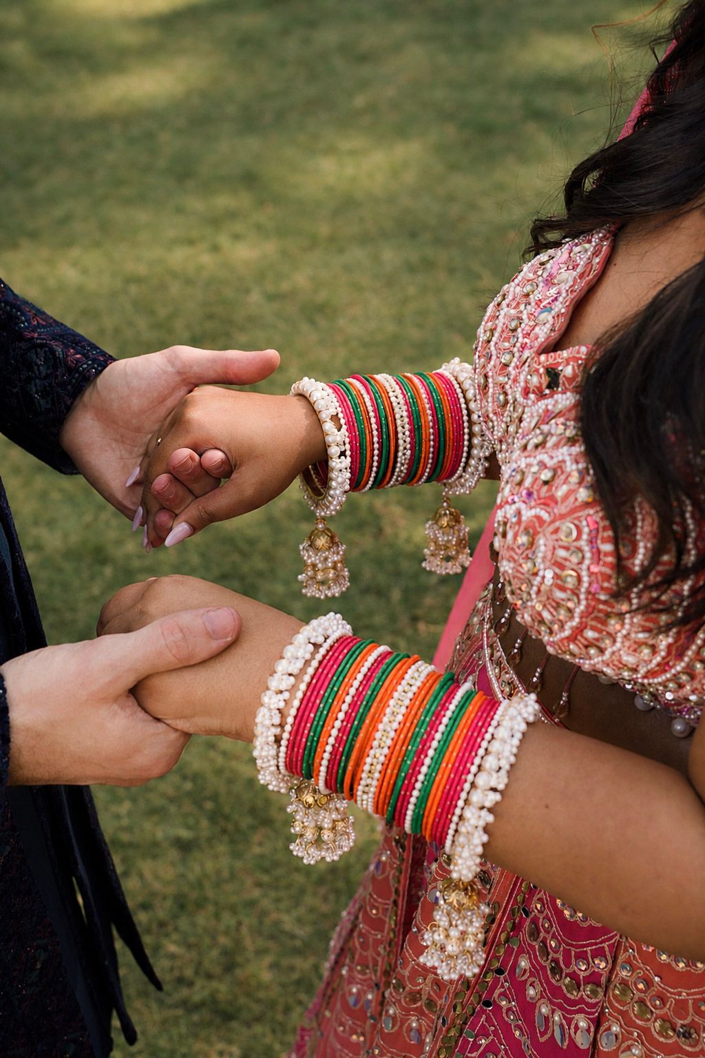 Indian bride in traditional red embroidered outfit with colorful bangles and jhumka earrings holding hands outdoors.