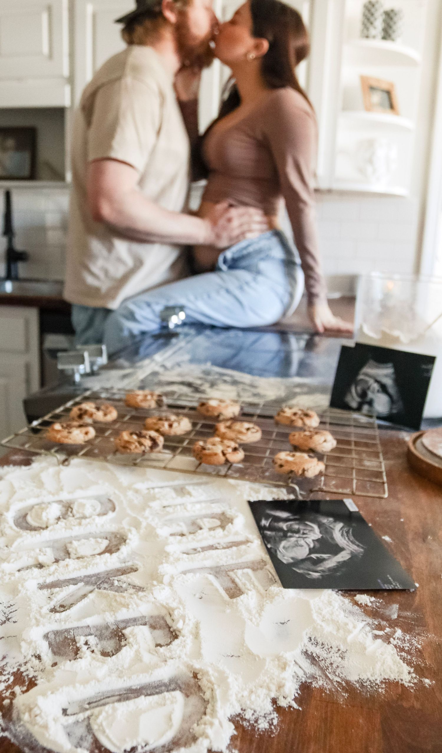A couple bakes cookies together in their kitchen with flour scattered on a wooden countertop and cooling racks nearby.