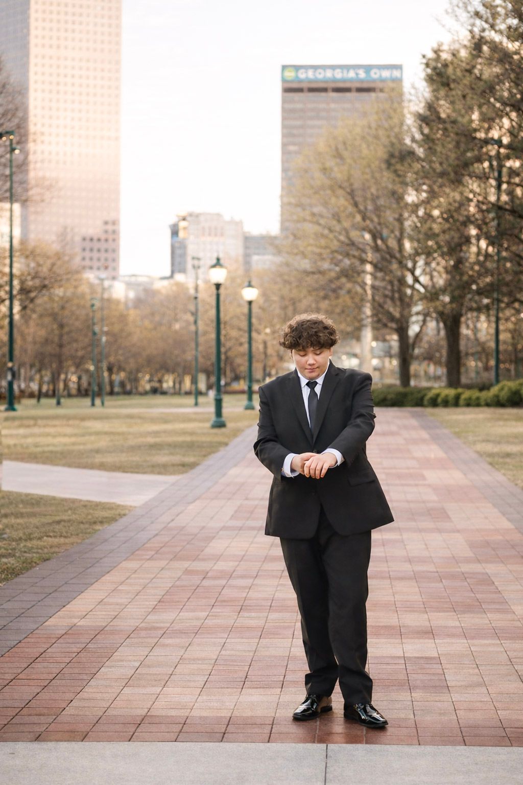 Young man in black suit checking his watch on a brick pathway in an urban park with city skyline backdrop.