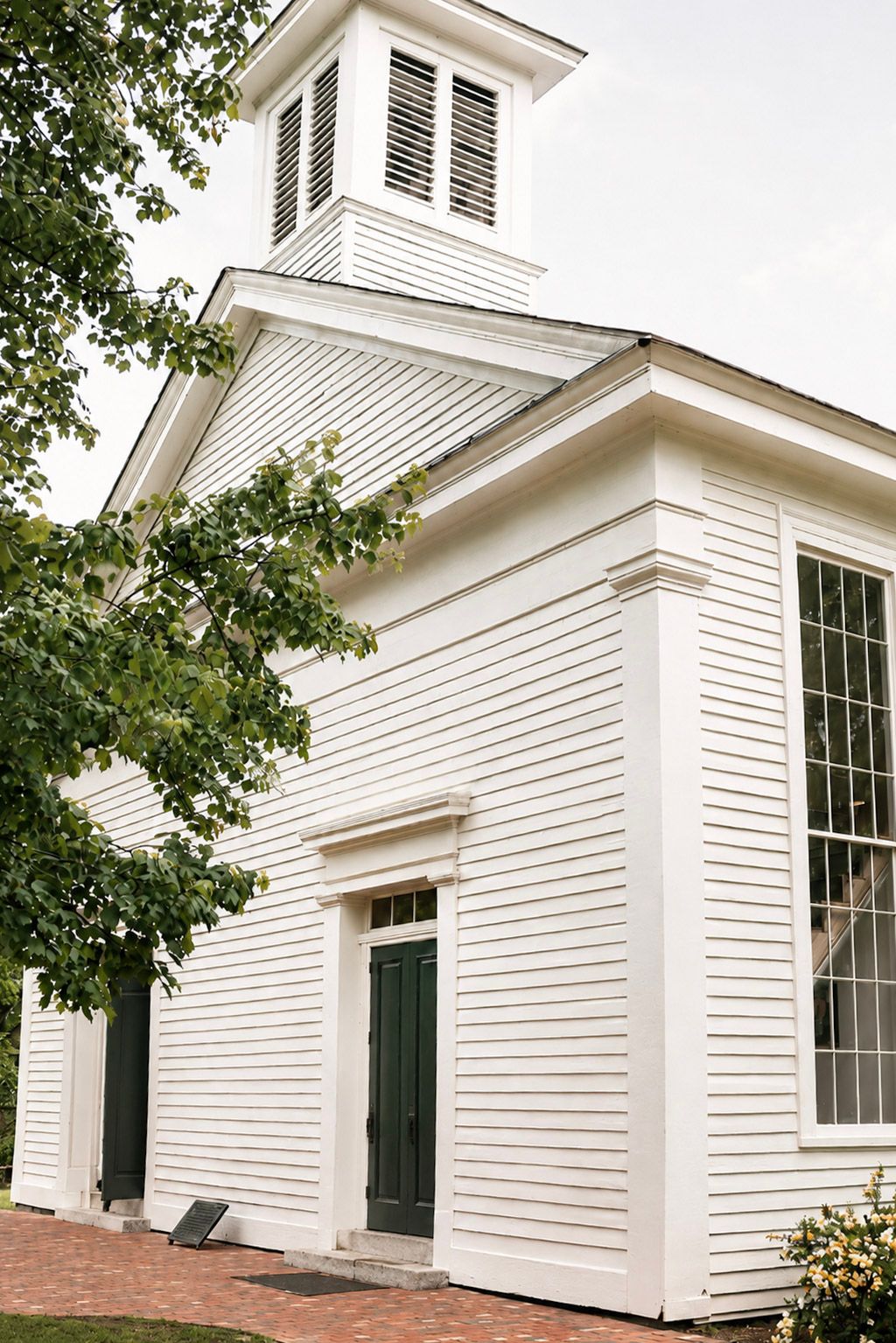 White colonial church with green door, cupola, and brick pathway surrounded by trees.